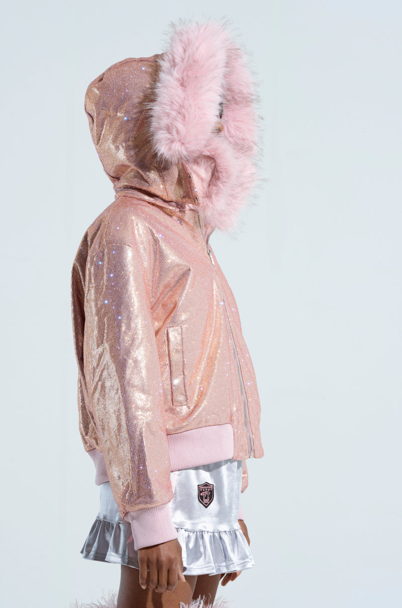 A person wears the EYES ON ME RHINESTONE PUFFER, a Barbie skiing outfit featuring a sparkly pink hooded jacket with fluffy fur trim and a shiny silver skirt. The head is turned to the side, and the arms rest by the sides against a plain light backdrop.