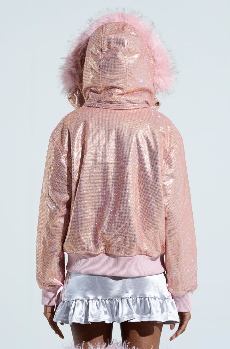 Rear view of a person wearing the EYES ON ME RHINESTONE PUFFER, a sparkly pink hooded jacket with fluffy pink trim, paired with a short, layered white skirt. The background is a plain light blue, reminiscent of a Barbie skiing outfit.