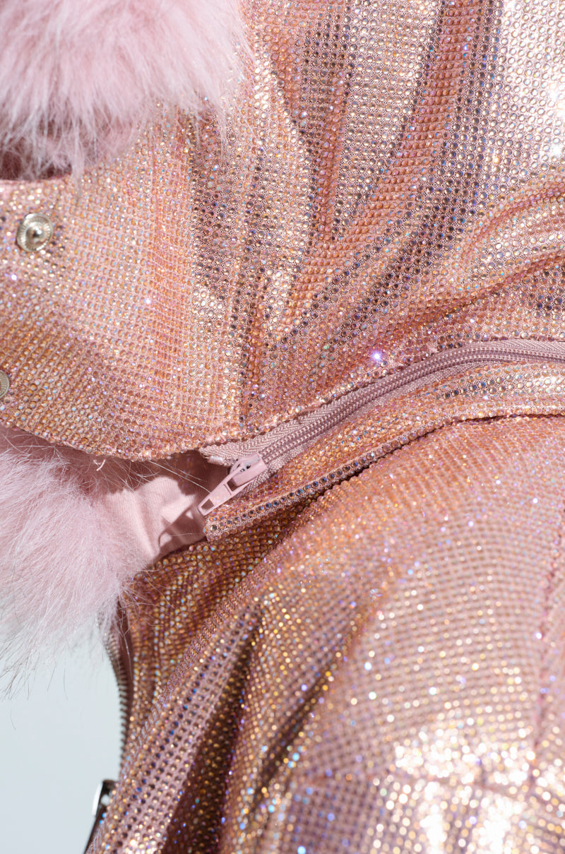A close-up view of the EYES ON ME RHINESTONE PUFFER showcases its pink, glittery fabric adorned with sequins and a fuzzy metallic fabrication zipper. The brilliantly reflective, textured surface catches the light beautifully. When partially unzipped, the zipper reveals an additional layer of soft, plush material underneath, reminiscent of this rhinestone puffer's cozy interior.