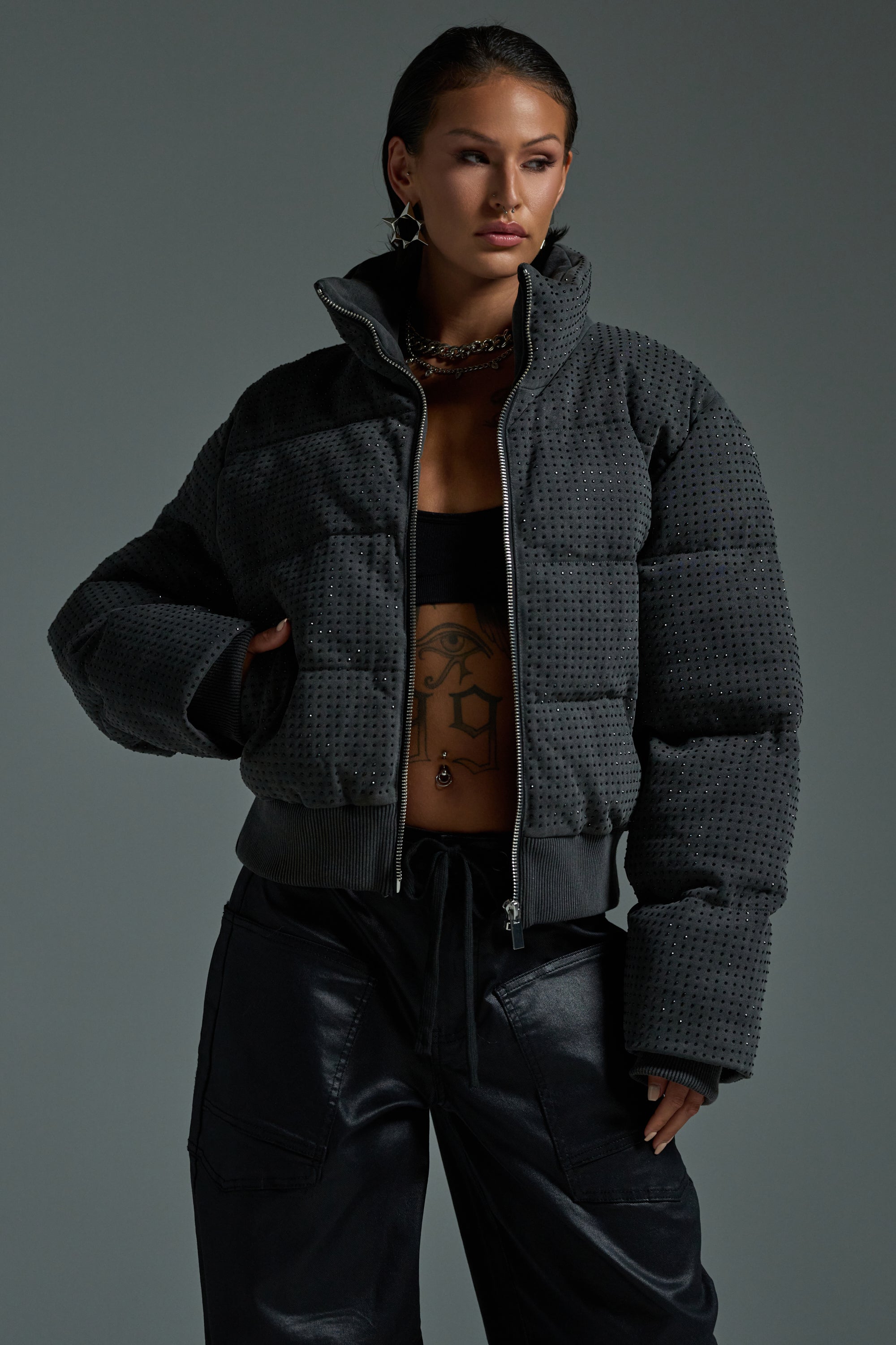 A woman poses confidently against a plain background in the HOUDINI RHINESTONE PUFFER over a black crop top, displaying her torso tattoo, styled with black cargo pants, earrings, and a choker.