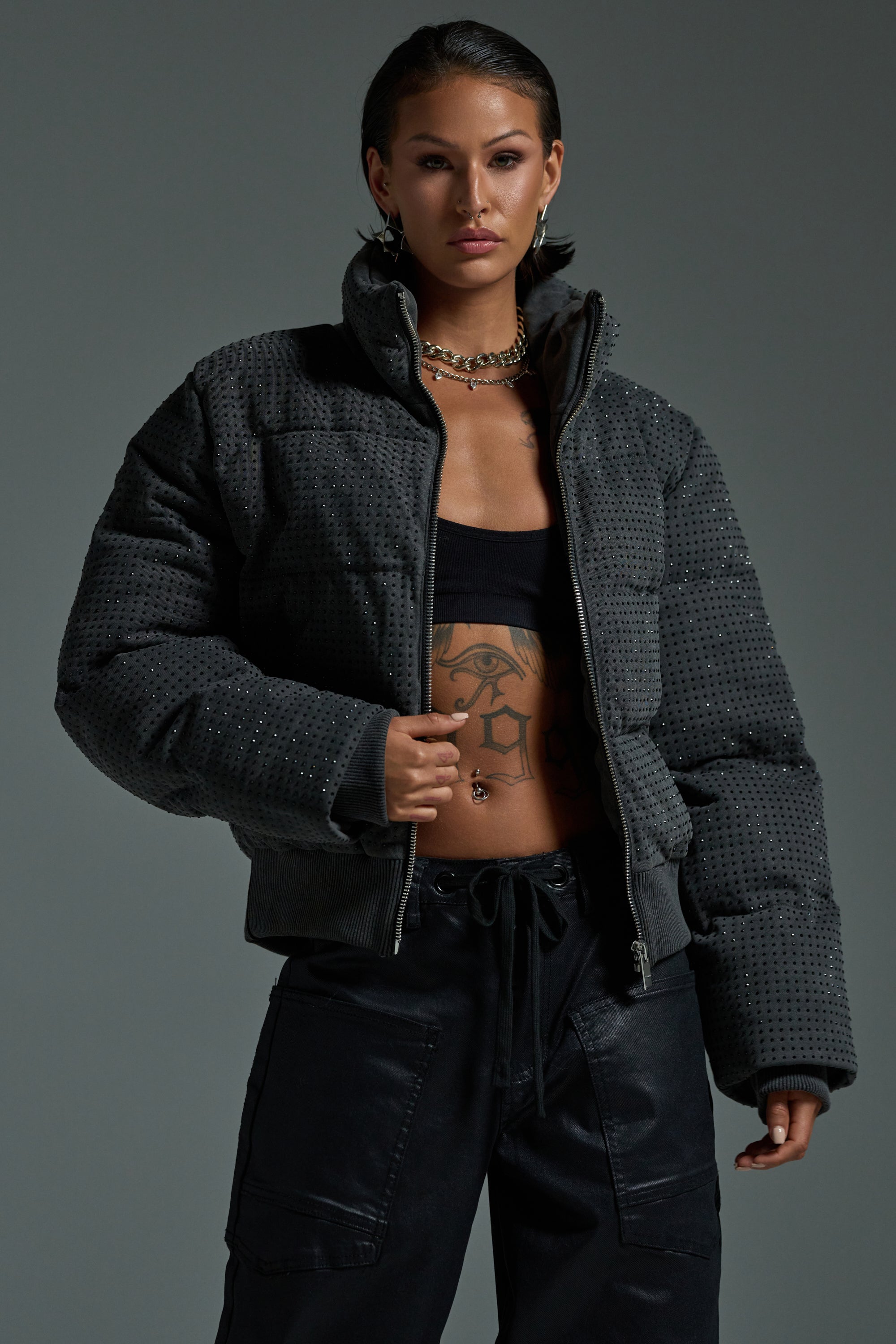 A woman poses confidently in the HOUDINI RHINESTONE PUFFER over a black crop top and cargo pants, accessorized with layered necklaces and showing visible tattoos, all set against a gray background.