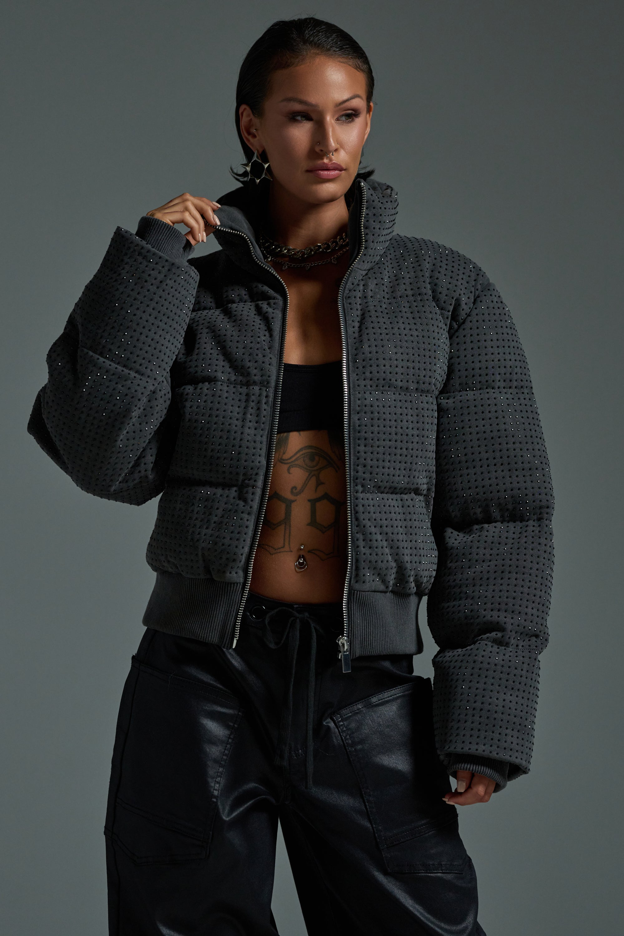 A woman stands confidently against a gray background, wearing the HOUDINI RHINESTONE PUFFER over a black crop top and pants. She accessorizes with layered necklaces and has her dark hair pulled back.