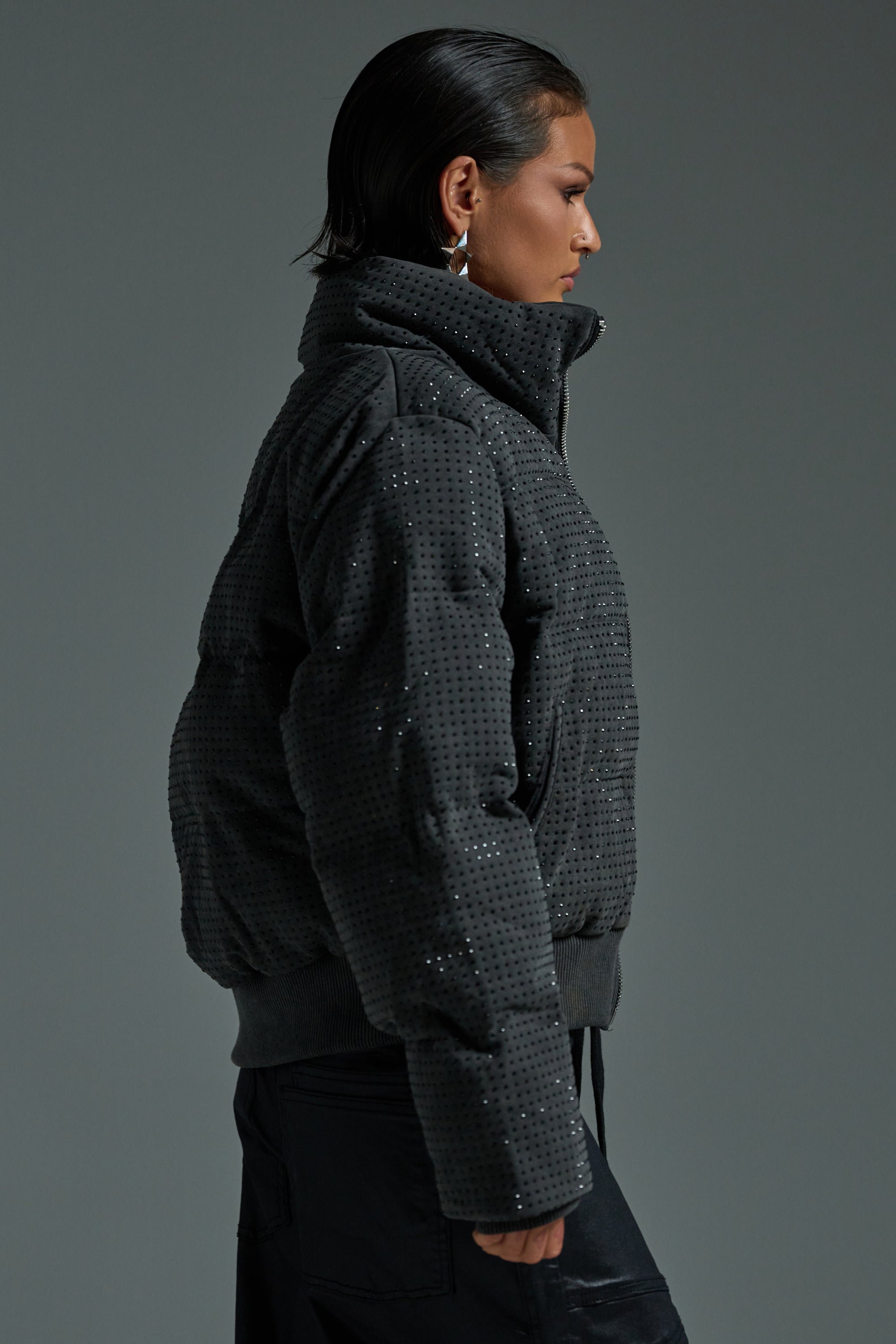 A person with slicked-back hair wears the HOUDINI RHINESTONE PUFFER and black pants, standing in profile against a plain gray background.