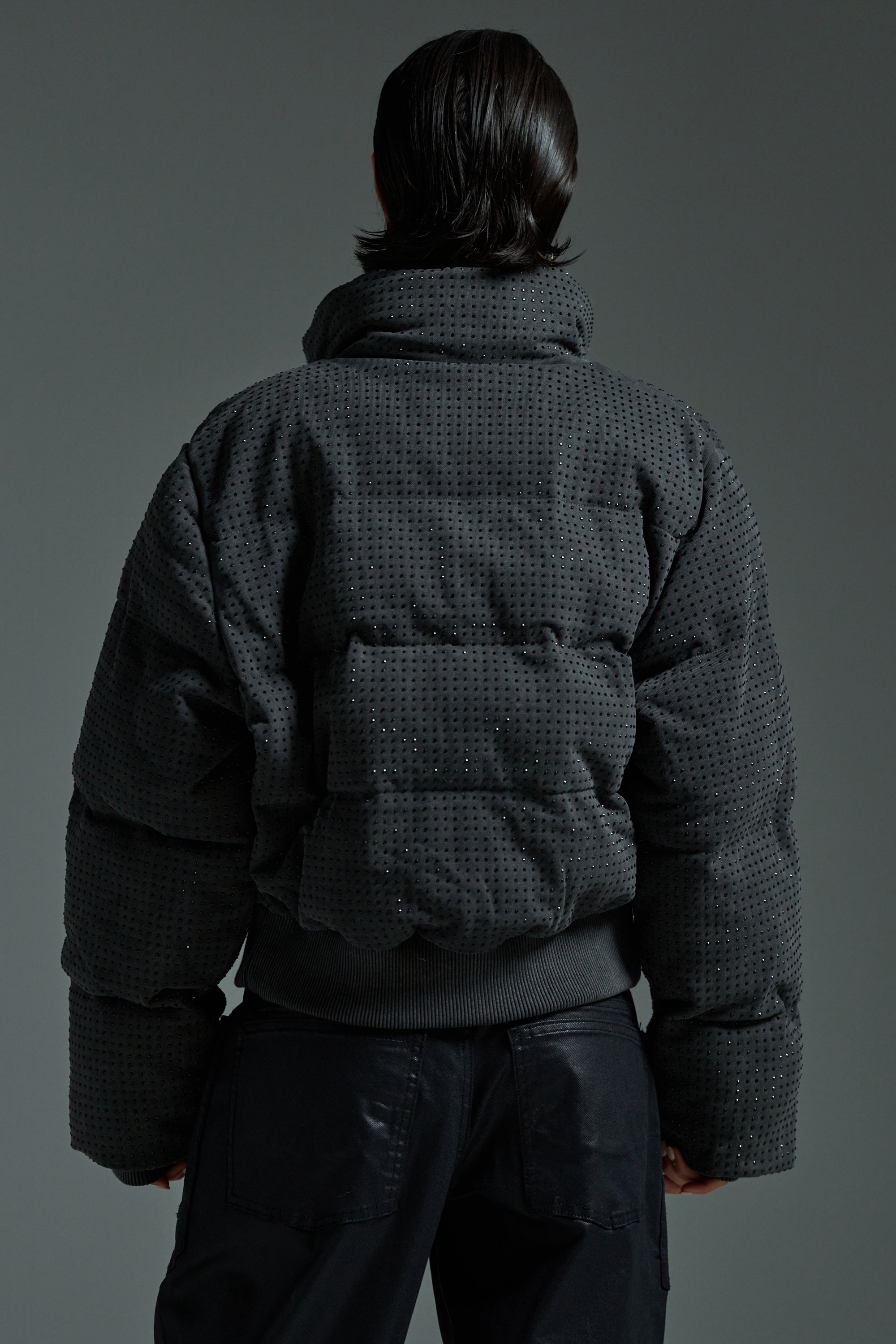 A person with dark, shoulder-length hair wears the HOUDINI RHINESTONE PUFFER and dark pants, seen from behind against a plain gray background.