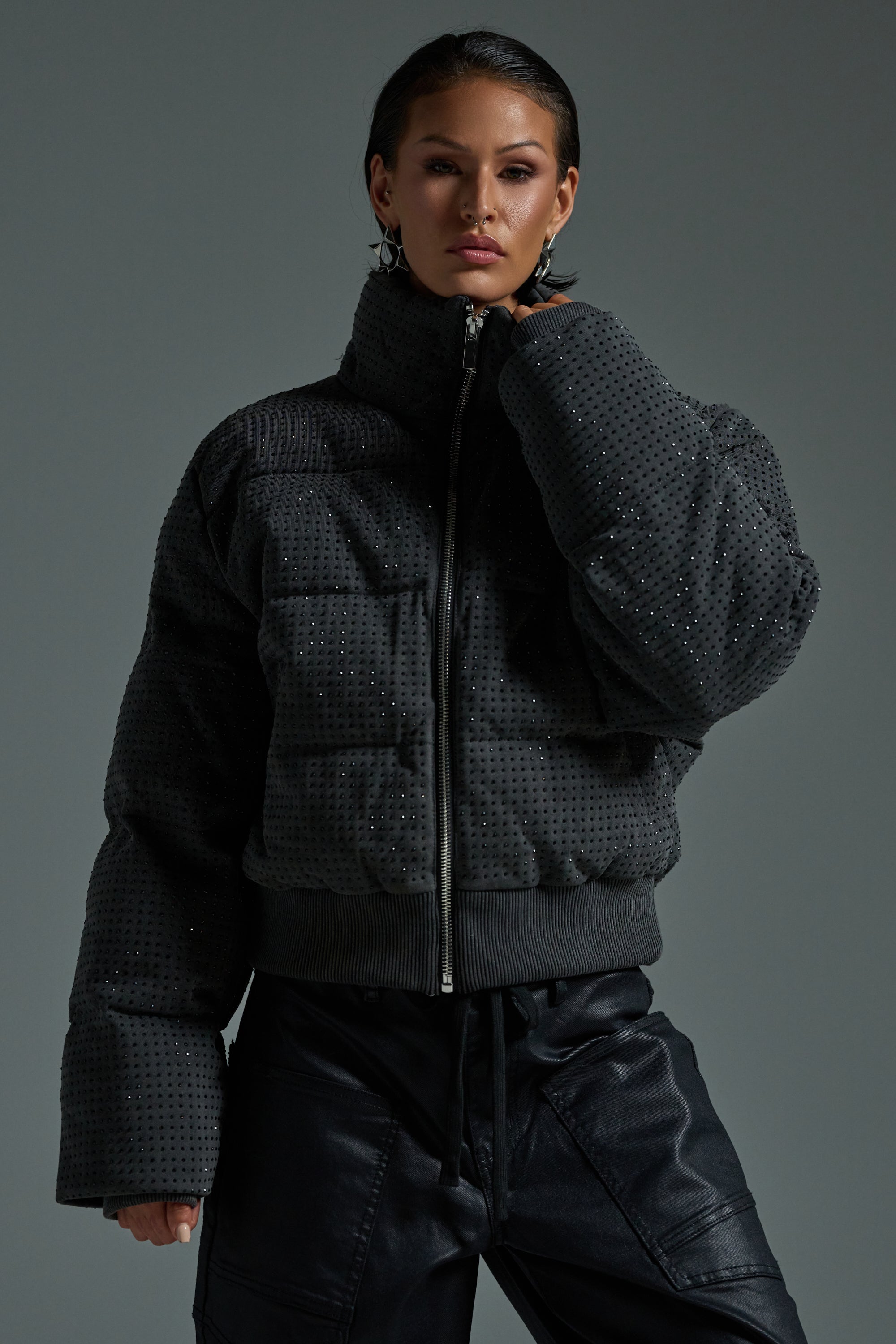 A person poses confidently in the HOUDINI RHINESTONE PUFFER—featuring a high collar and wide sleeves—paired with black leather pants, against a plain gray background.