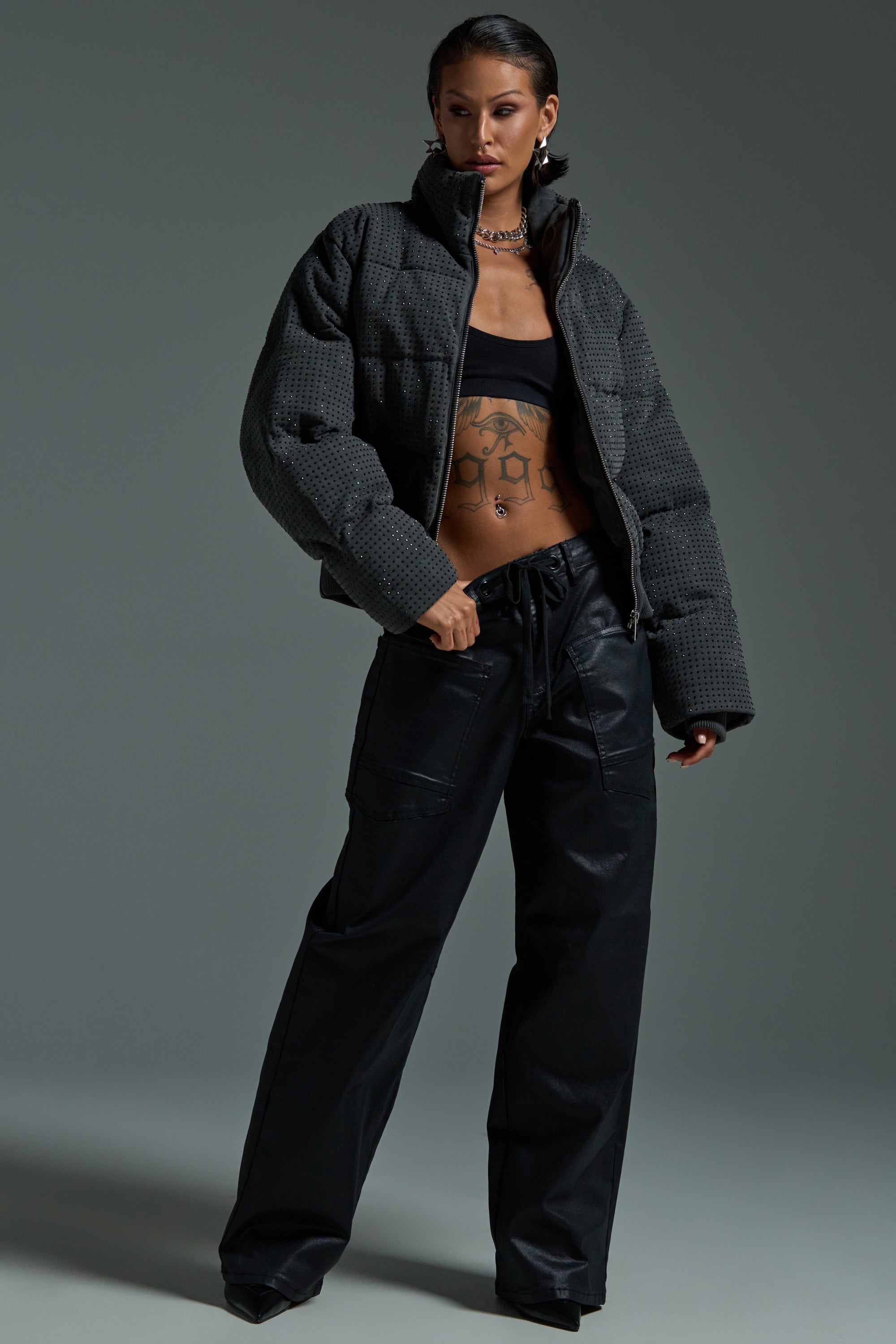 A person poses confidently in the HOUDINI RHINESTONE PUFFER, black bandeau top, loose cargo pants, and chunky jewelry, showing off torso tattoos against a plain gray background.