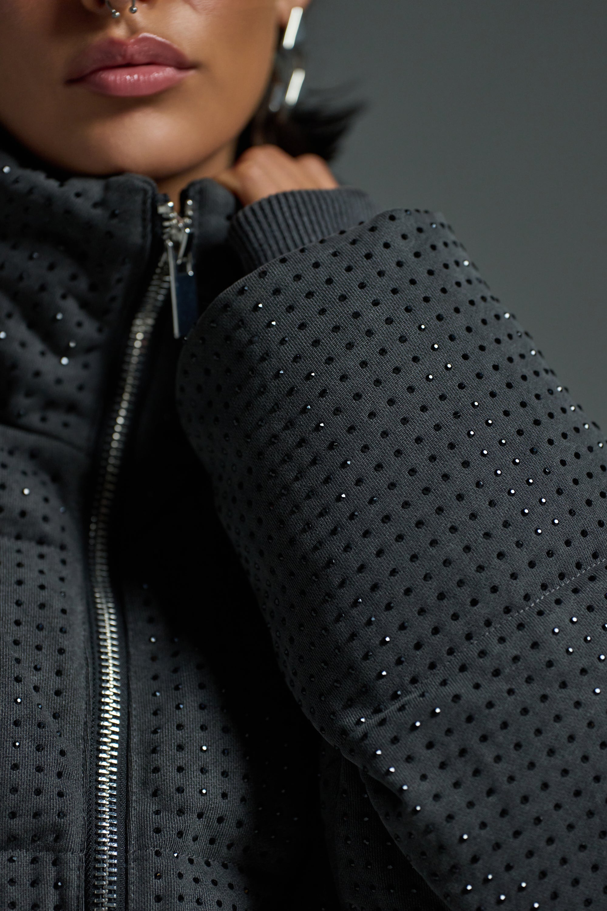 A close-up shows a person wearing the HOUDINI RHINESTONE PUFFER, a dark, sparkling jacket with stud accents and a zipper. Only the lower half of their face is visible, highlighting their septum piercing and hoop earrings.