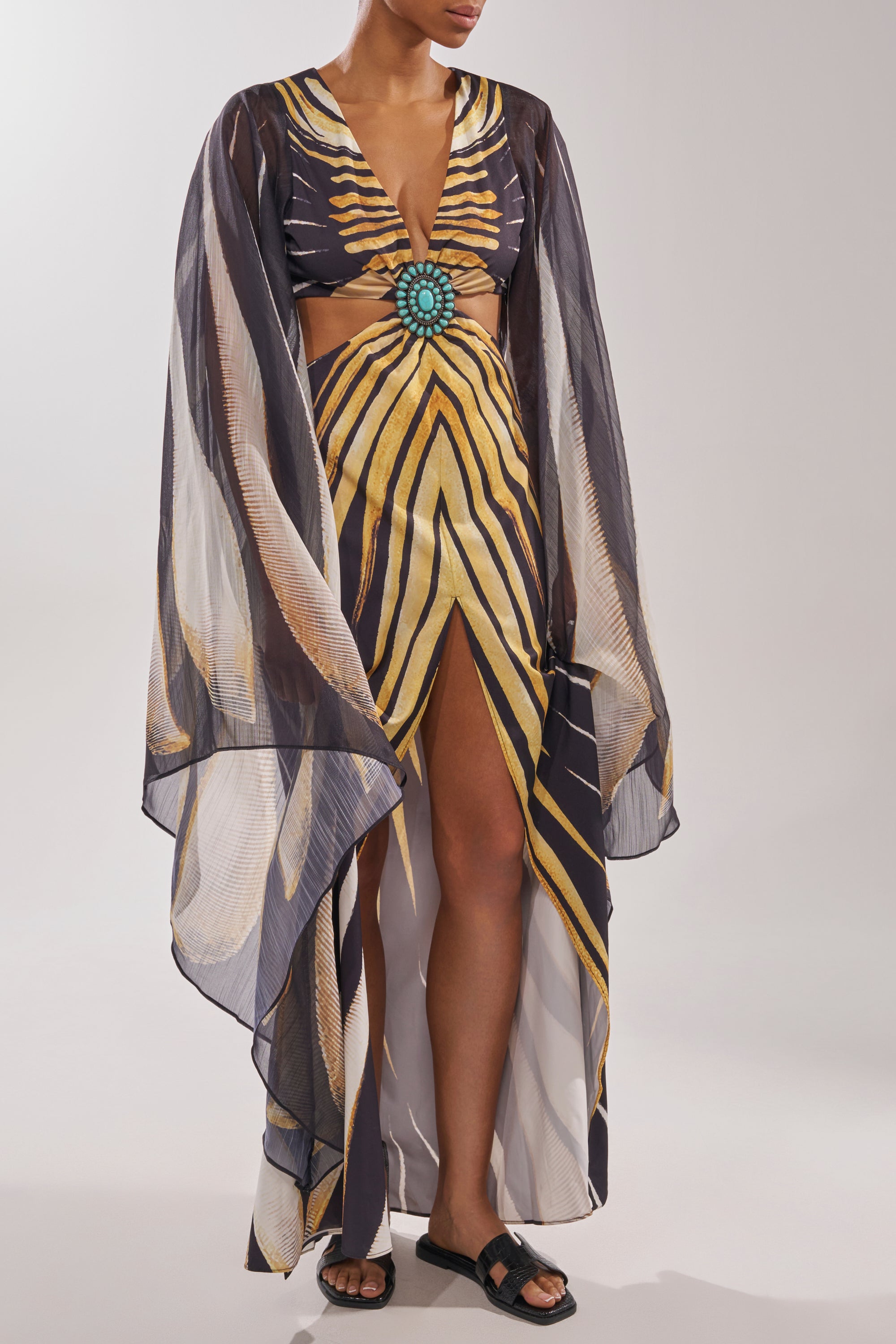 A woman wears a flowing sheer dress with yellow and black stripes, wide sleeves, a turquoise brooch, waist cutout, and front slit. She pairs it with AKIRA HOWDY BLACK CROC SLIP ON FLAT SANDAL. Only her lower face is visible. 1