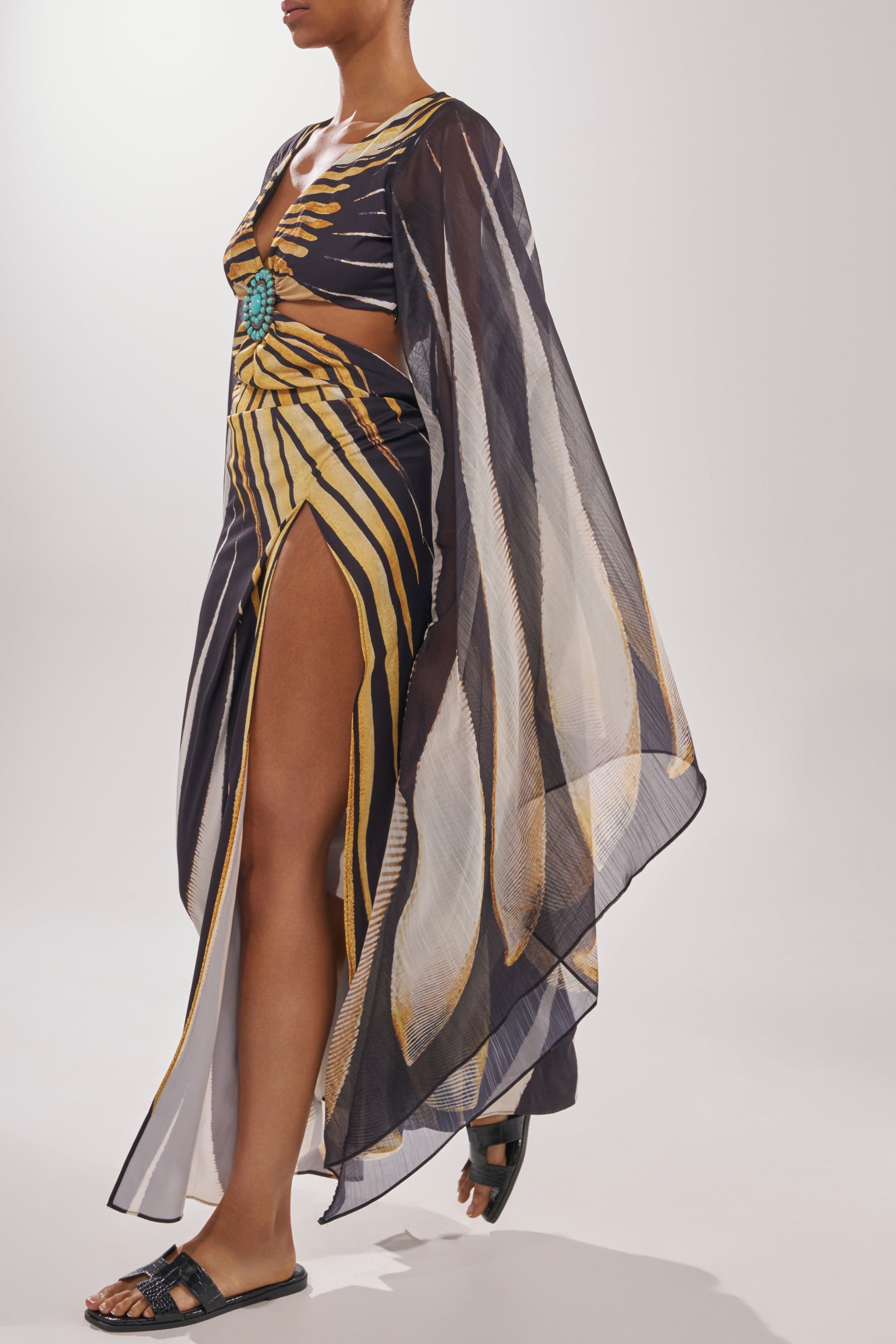 A person in a sheer black and yellow cutout dress with a turquoise center detail and high slit, paired with AKIRA HOWDY BLACK CROC SLIP ON FLAT SANDALS in crocodile-embossed faux leather. 2