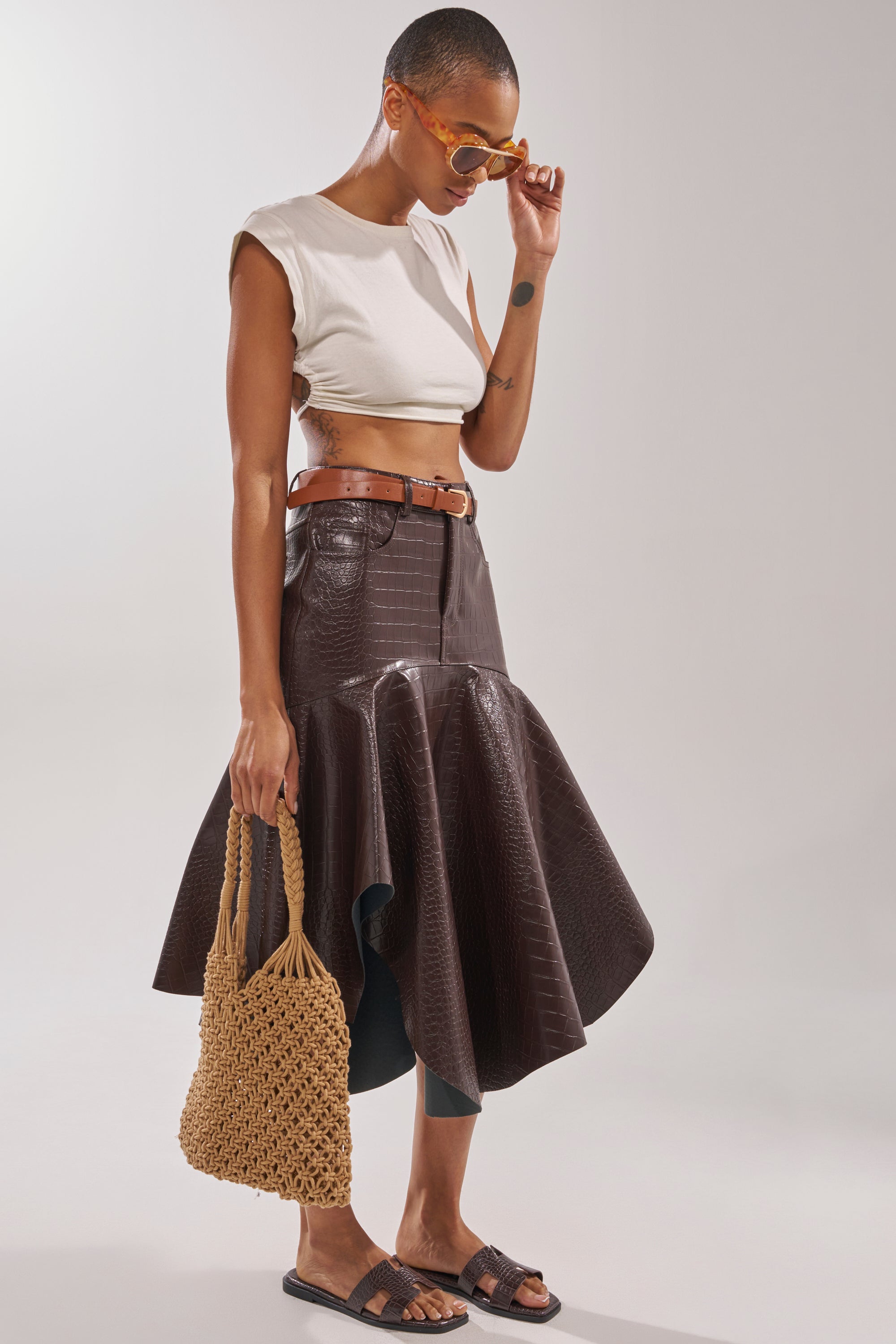 A person in a white crop top, brown crocodile-embossed faux leather flared skirt, belt, dark sunglasses, and AKIRA HOWDY BROWN CROC SLIP ON FLAT SANDAL, holding a woven handbag and standing against a neutral background. 3