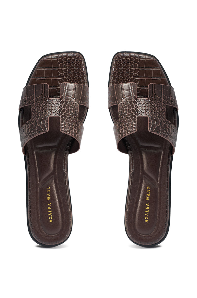 The AKIRA HOWDY BROWN CROC SLIP ON FLAT SANDAL features brown crocodile-embossed faux leather, square toes, and wide crisscross straps. "AZALEA WANG" is printed in gold on the insoles. The sandals are shown from above on a white background. 5