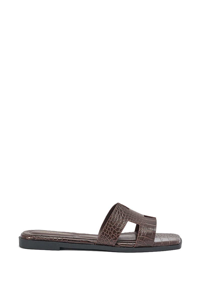 The AKIRA HOWDY BROWN CROC SLIP ON FLAT SANDAL features a flat sole, wide crocodile-embossed faux leather strap with side cutouts, and a chic square-toe design, shown on a white background. 6