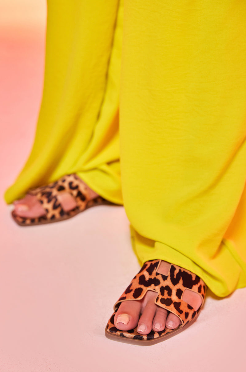 A person wearing bright yellow pants stands on a light pink surface, showing just their toes in the AKIRA HOWDY FAUX SUEDE LEOPARD SLIP ON SANDAL with a comfort cushion footbed.