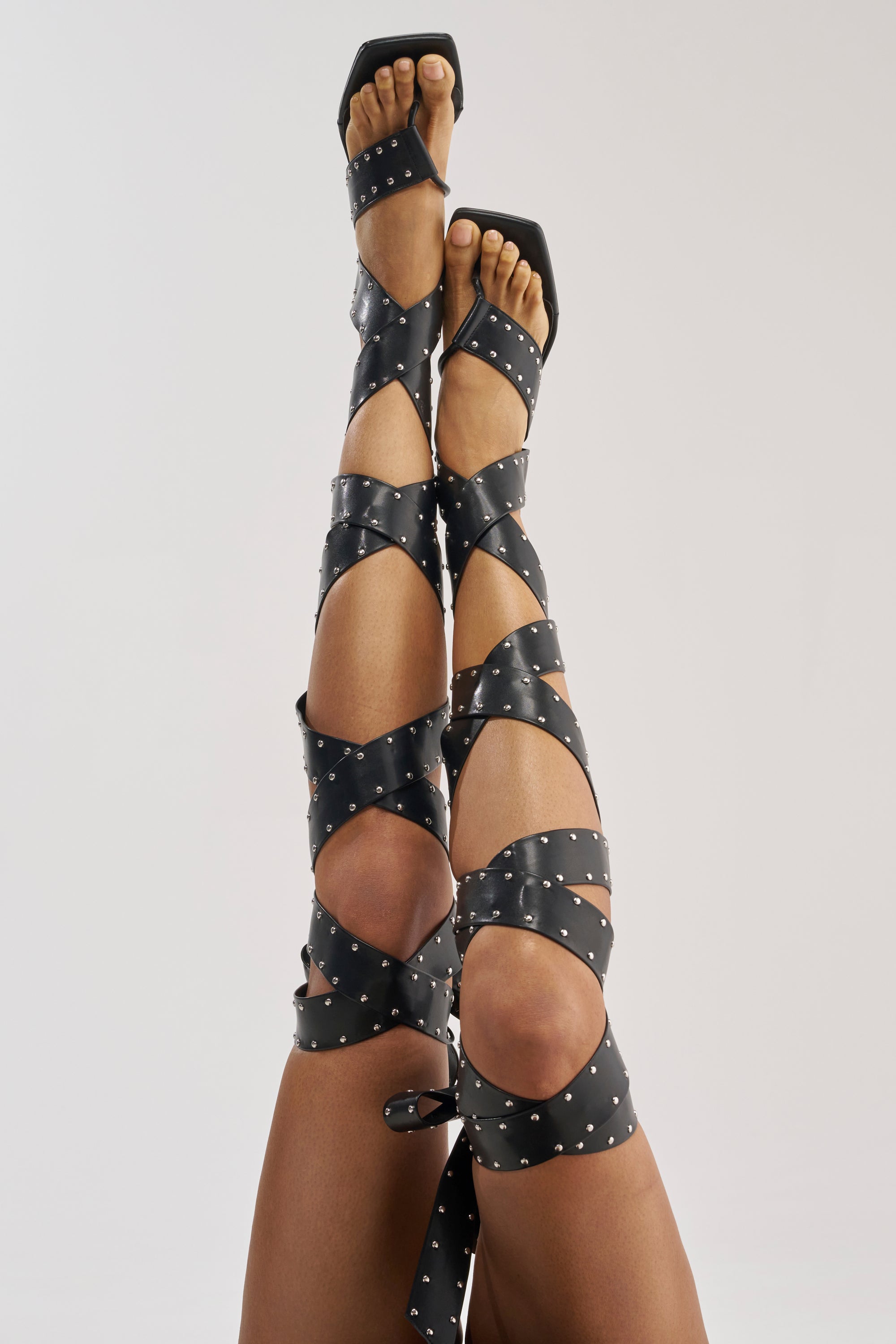 A person’s legs are raised and crossed, showcasing the AZALEA WANG HOWLITE BLACK STUDDED STRAPPED SANDAL—black faux leather stiletto heels with long, studded straps that lace up to the knees—standing out strikingly against the plain, light-colored background.