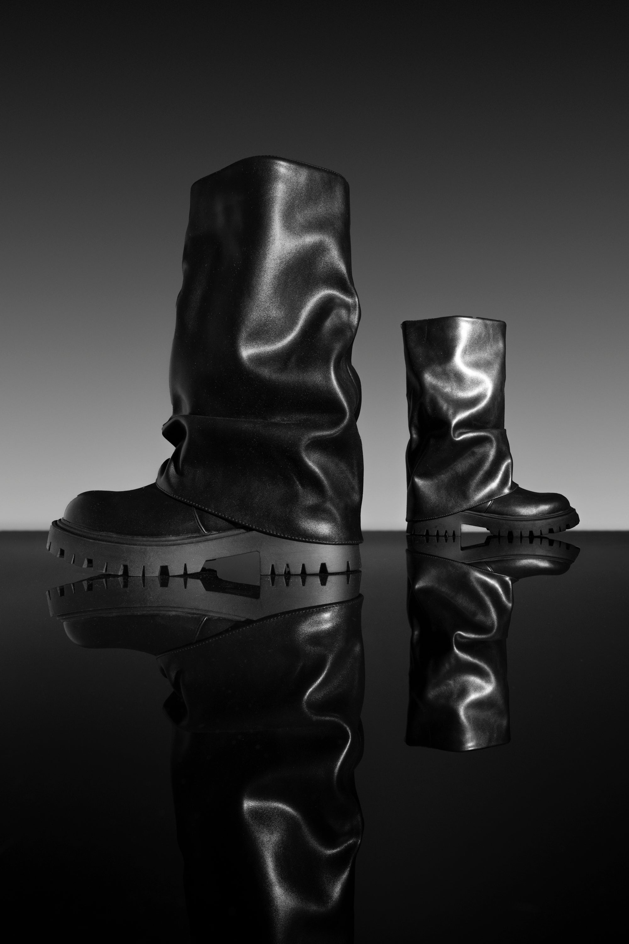 Two AZALEA WANG HOYT BLACK FLAT RUCHED BOOTIE boots in black faux leather with chunky soles stand on a reflective surface, creating a mirrored effect. The background fades from dark to light, highlighting the boots’ smooth texture and their slouched, foldover shaft shape.