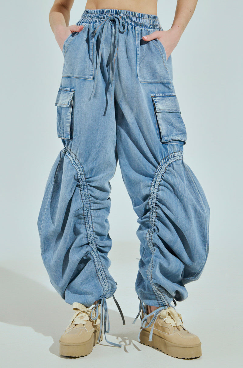 A person is wearing high-rise, relaxed fit MIA FLOW PANT denim cargo pants in light blue with customizable pantlegs and drawstring details for a balloon effect, complemented by multiple pockets. Beige chunky sneakers with cream laces complete the look.