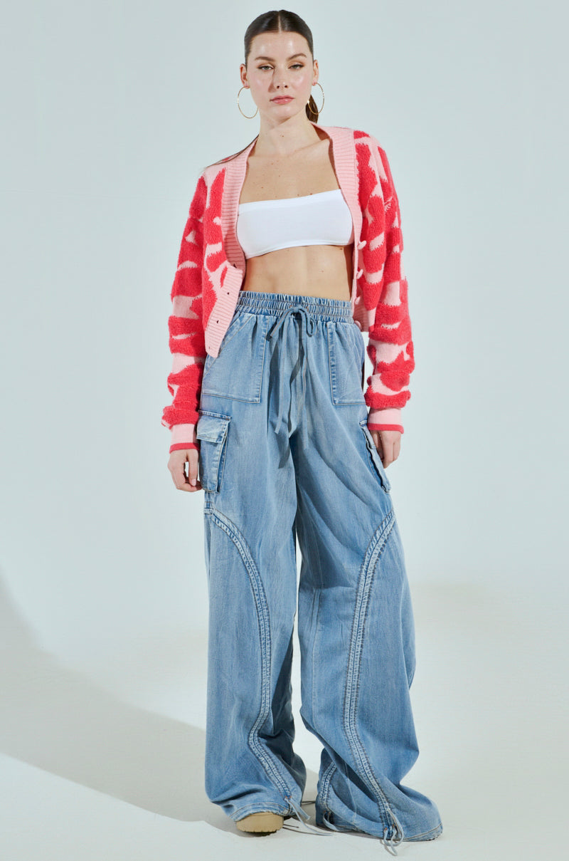 A person stands against a simple background, wearing a pink and red patterned cardigan, a white crop top, MIA FLOW PANT high-rise relaxed fit denim-inspired drawstring pants. The outfit is completed with hoop earrings and beige shoes.