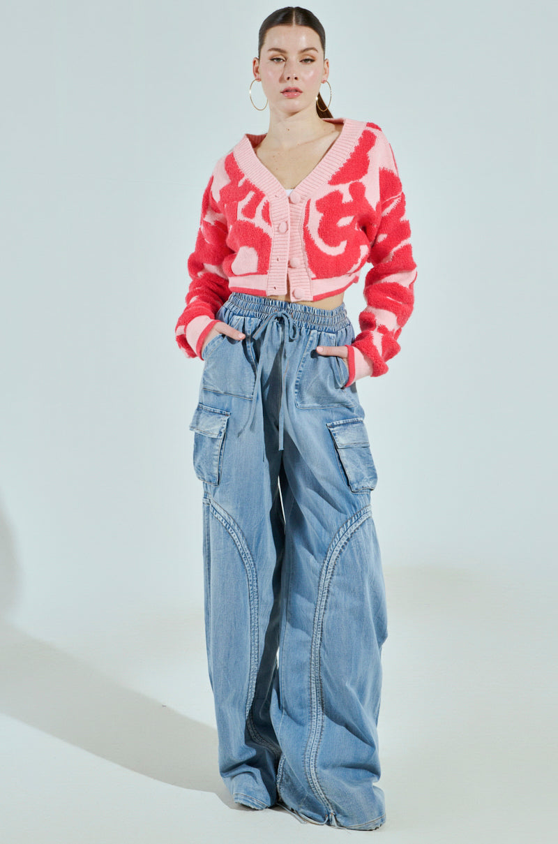 The individual stands against a light background wearing a pink and white patterned cropped cardigan with the high-waisted MIA FLOW PANT, designed with a relaxed, denim-inspired high-rise fit. They accessorize with hoop earrings and have their hair pulled back for a touch of effortless elegance.