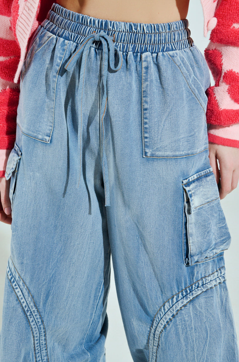 Cropped image of a person wearing the MIA FLOW PANT, featuring light blue denim-inspired high-rise relaxed fit, large pockets, and an elastic drawstring waistband. They are also sporting a pink and red patterned jacket.