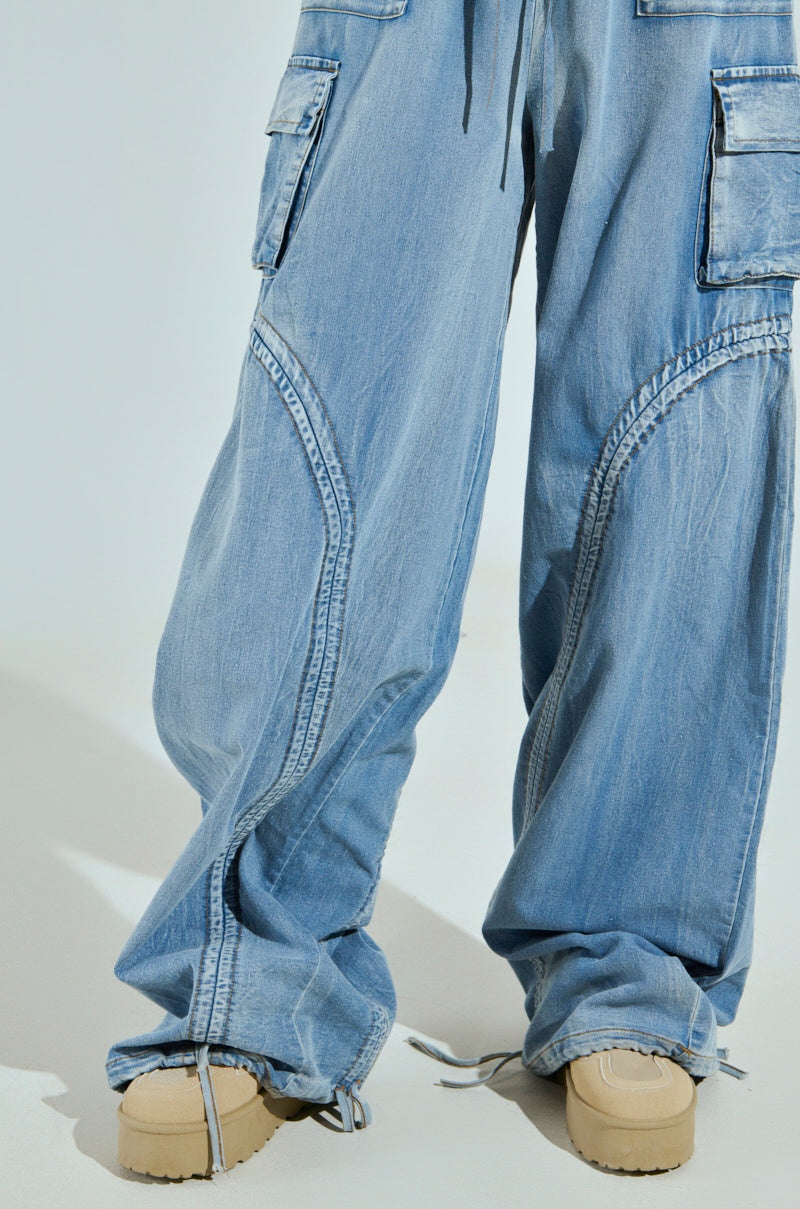 Someone is wearing the MIA FLOW PANT in light blue, featuring high-rise relaxed fit and cargo style with multiple pockets, paired with tan shoes that have thick soles. The pant legs crumple slightly above the shoes against a plain background.