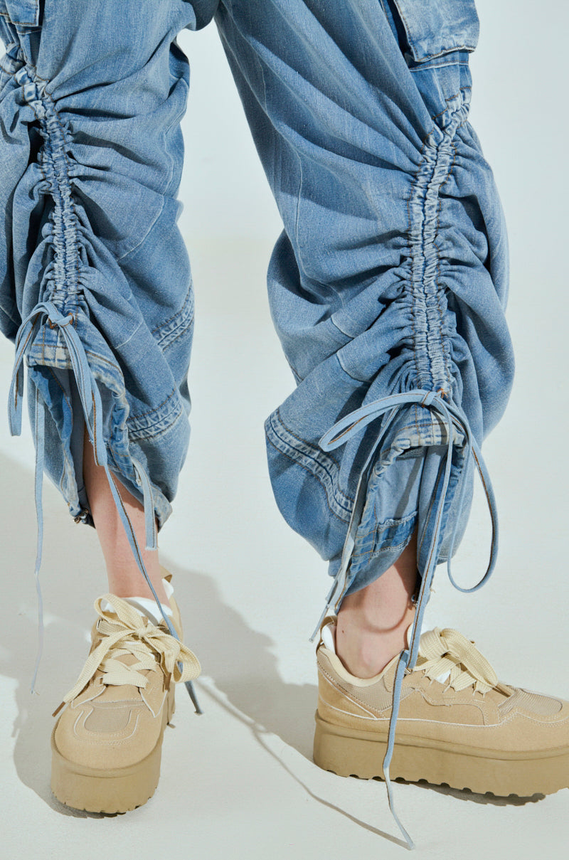 A person stands on a light background wearing MIA FLOW PANT, high-rise, relaxed fit in ruched blue denim-inspired fabric with string-tied gathered details, paired with beige sneakers featuring thick soles and large laces.
