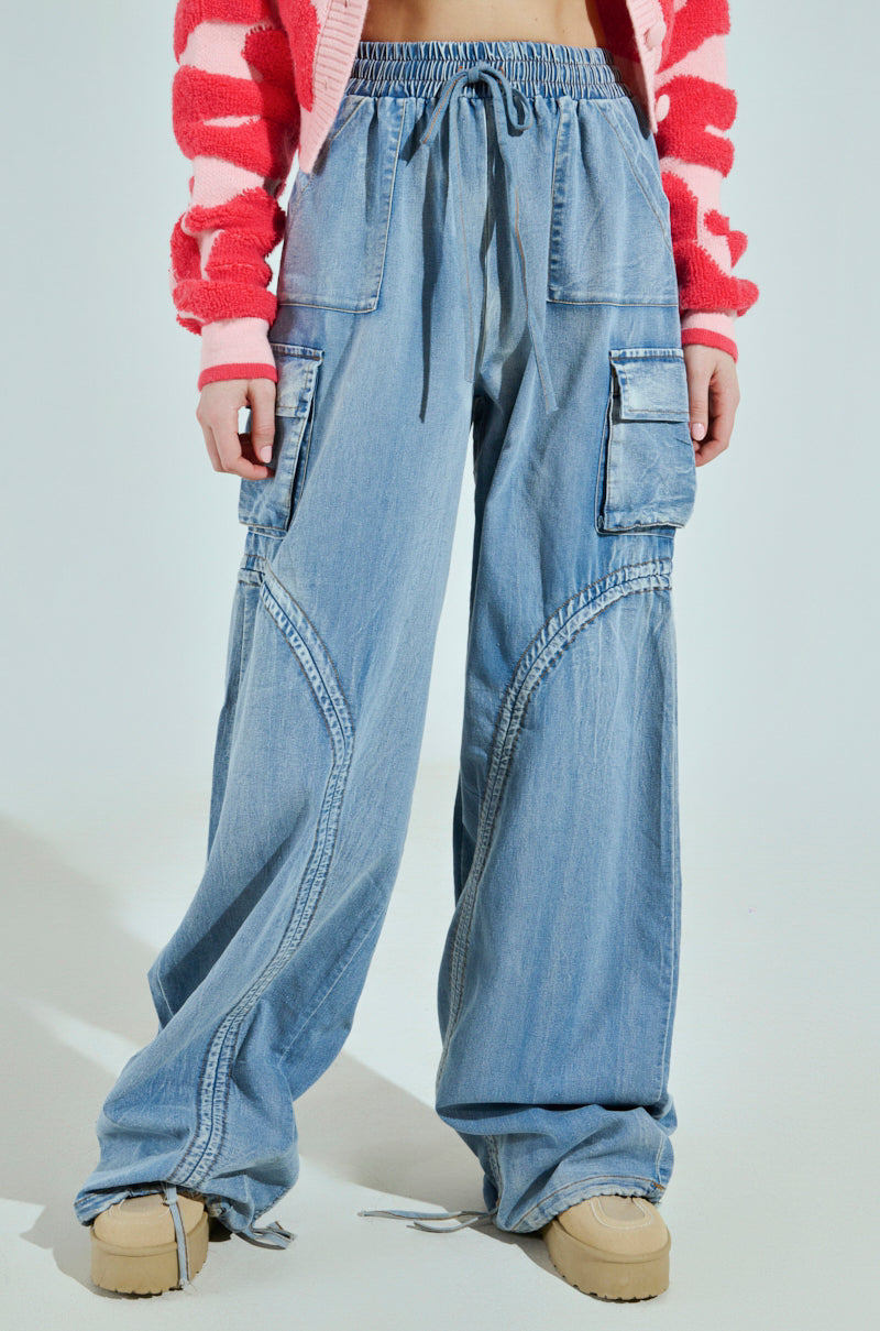 The person is wearing MIA FLOW PANT—light blue, high-rise relaxed fit cargo pants with denim-inspired fabric, large pockets, and customizable pantlegs. They also sport a red and pink striped sweater and beige platform shoes against a plain, light-colored background.