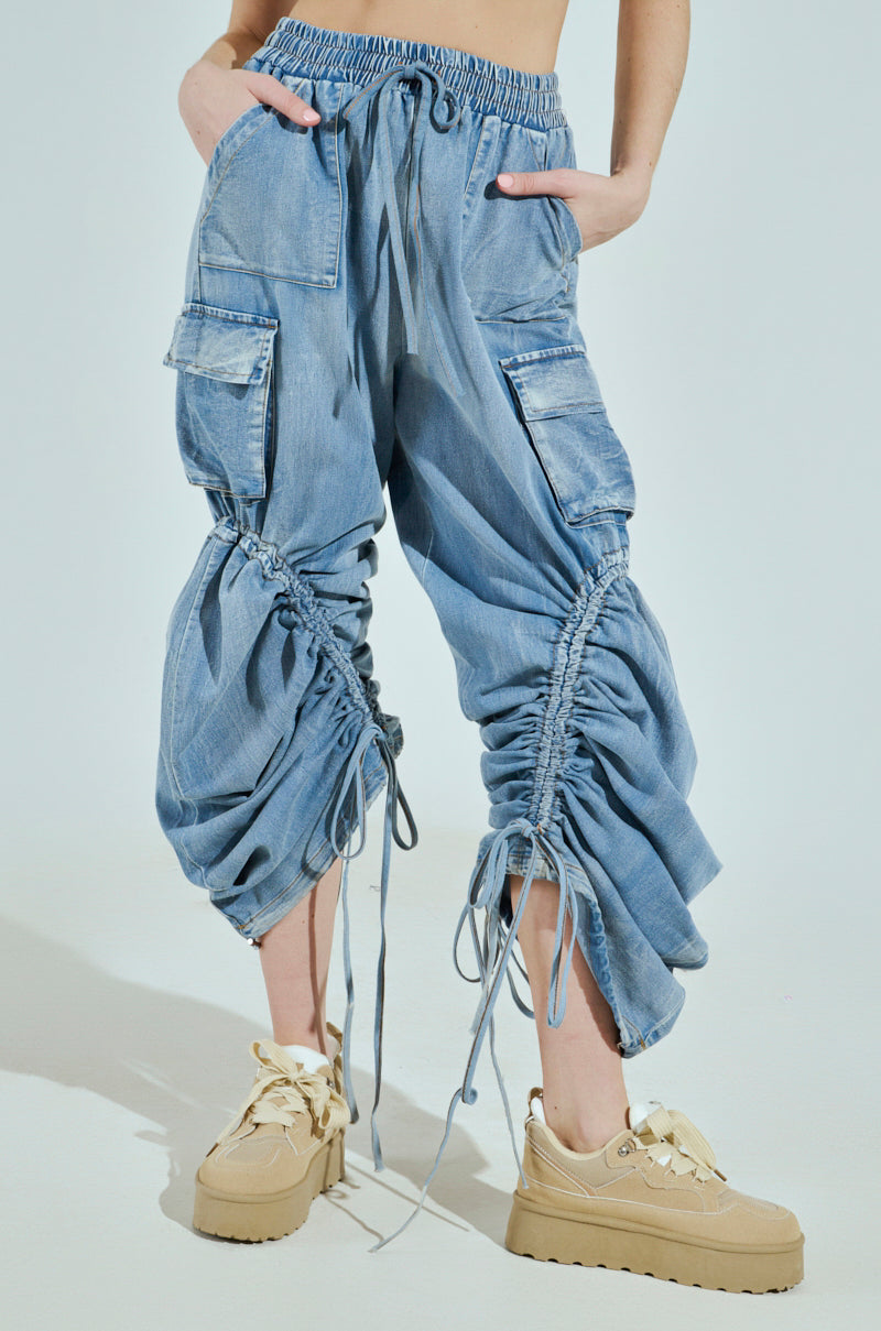 A person is wearing the MIA FLOW PANT, loose and light blue denim cargo pants featuring ruched detailing and drawstrings at the knees for customizable pantlegs, paired with beige chunky sneakers. Hands in pockets highlight a casual and trendy fashion style.