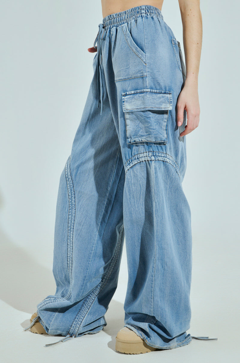 Someone is wearing the MIA FLOW PANT, which are loose-fitting light blue cargo jeans with customizable pantlegs and a drawstring waist. Featuring large side pockets and a slightly faded texture, these high-rise relaxed fit pants pair perfectly with beige shoes against a plain white background.