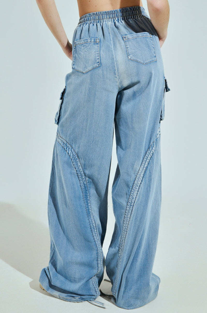 Someone is wearing the MIA FLOW PANT, featuring light blue, high-rise relaxed fit with denim-inspired fabric. These wide-leg pants include visible stitching, an elastic waistband, cargo-style side pockets, and two back pockets against a plain background.