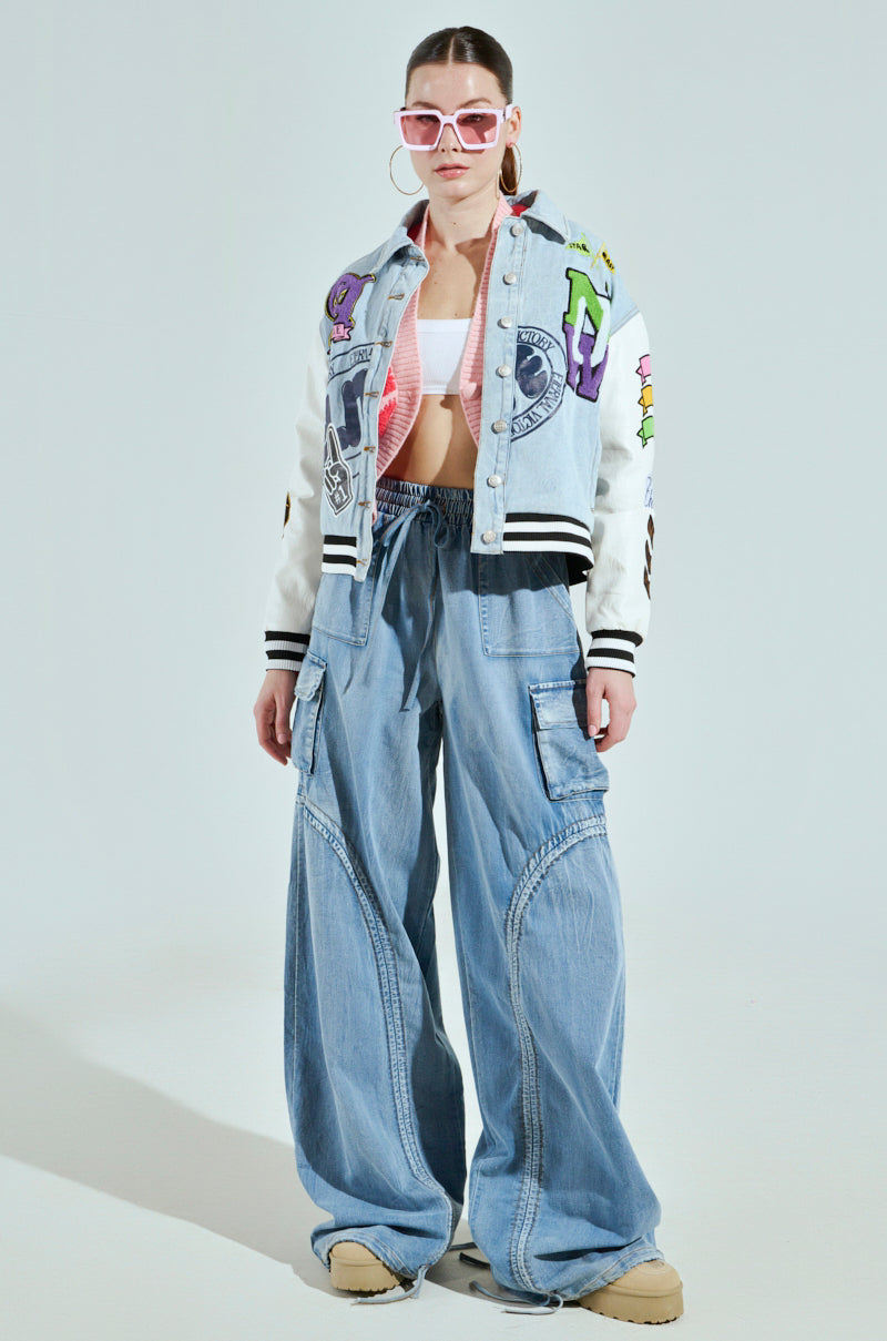 Wearing oversized, high-rise MIA FLOW PANTs and a colorful graphic bomber jacket over a white crop top, they confidently sport pink sunglasses and hoop earrings. Their sleek updo pairs perfectly with denim-inspired fabric and beige shoes.