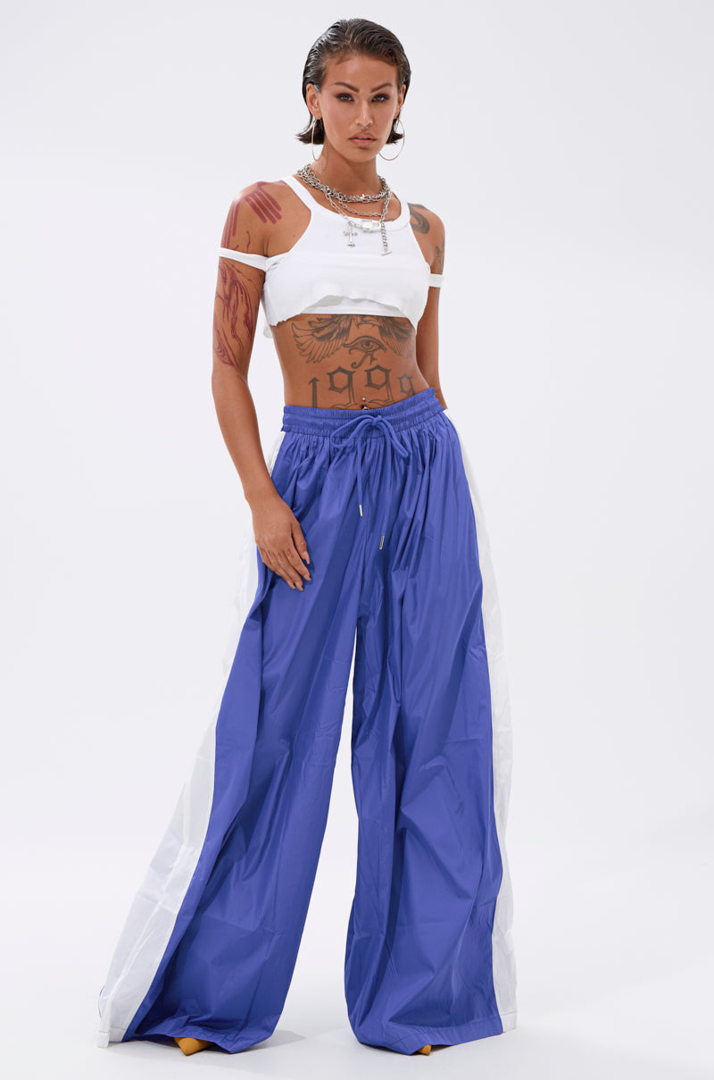 A person with wet, slicked-back hair wears a white crop top and the BREAKING THE RULES TRACK PANT IN ROYAL BLUE, featuring loose, high-rise fit and white side panels. Tattoos and layered necklaces add edge to this athleisure vibe. 0