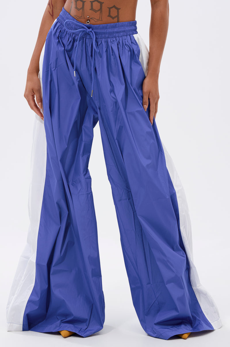 A person wears BREAKING THE RULES TRACK PANT IN ROYAL BLUE with white side panels and a drawstring waist, styled high-rise to reveal a tattooed midriff against a plain white background. 1