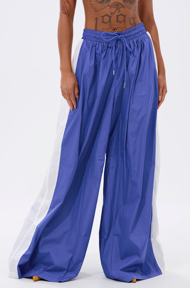 A person wears the BREAKING THE RULES TRACK PANT IN ROYAL BLUE, featuring a high-rise fit, white side panels, and drawstring waist. The athleisure look stands out against a plain background; tattoos are partially visible on the torso. 2