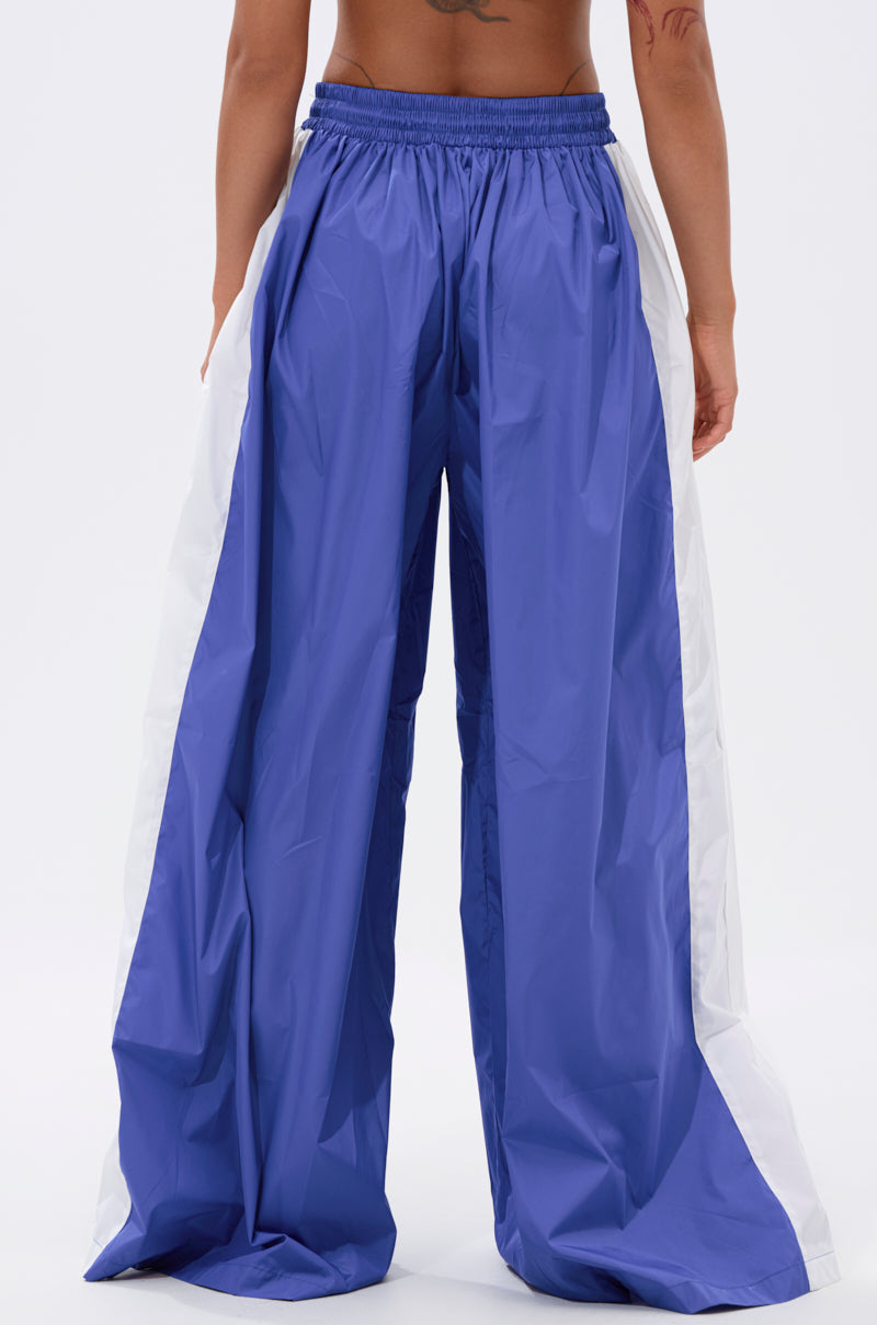 A person faces away, wearing BREAKING THE RULES TRACK PANT IN ROYAL BLUE—loose-fitting, high-rise track pants with wide white side panels, an elastic waistband, and shiny lightweight fabric. The background is plain white. 4