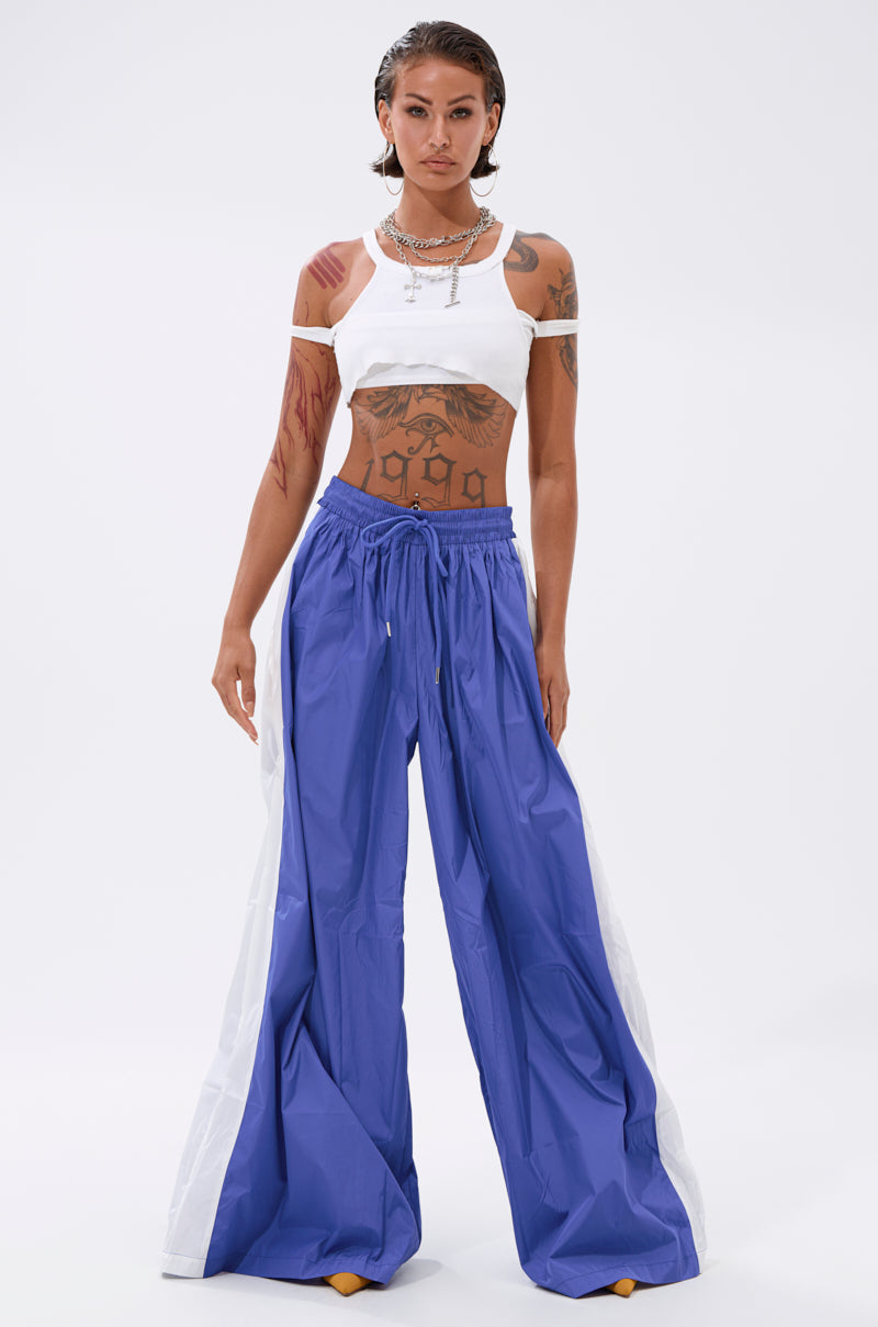 A person stands confidently against a white background, wearing a white cropped tank top and the BREAKING THE RULES TRACK PANT IN ROYAL BLUE with white side panels, styled with wet-look hair, visible tattoos, layered necklaces, and an athleisure vibe. 5