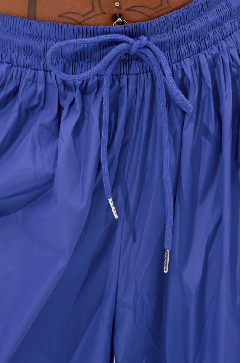 Close-up of a person wearing the BREAKING THE RULES TRACK PANT IN ROYAL BLUE, featuring a shiny finish and tied high-rise waistband. A portion of a tattoo and belly button piercing appear above the waist. 6
