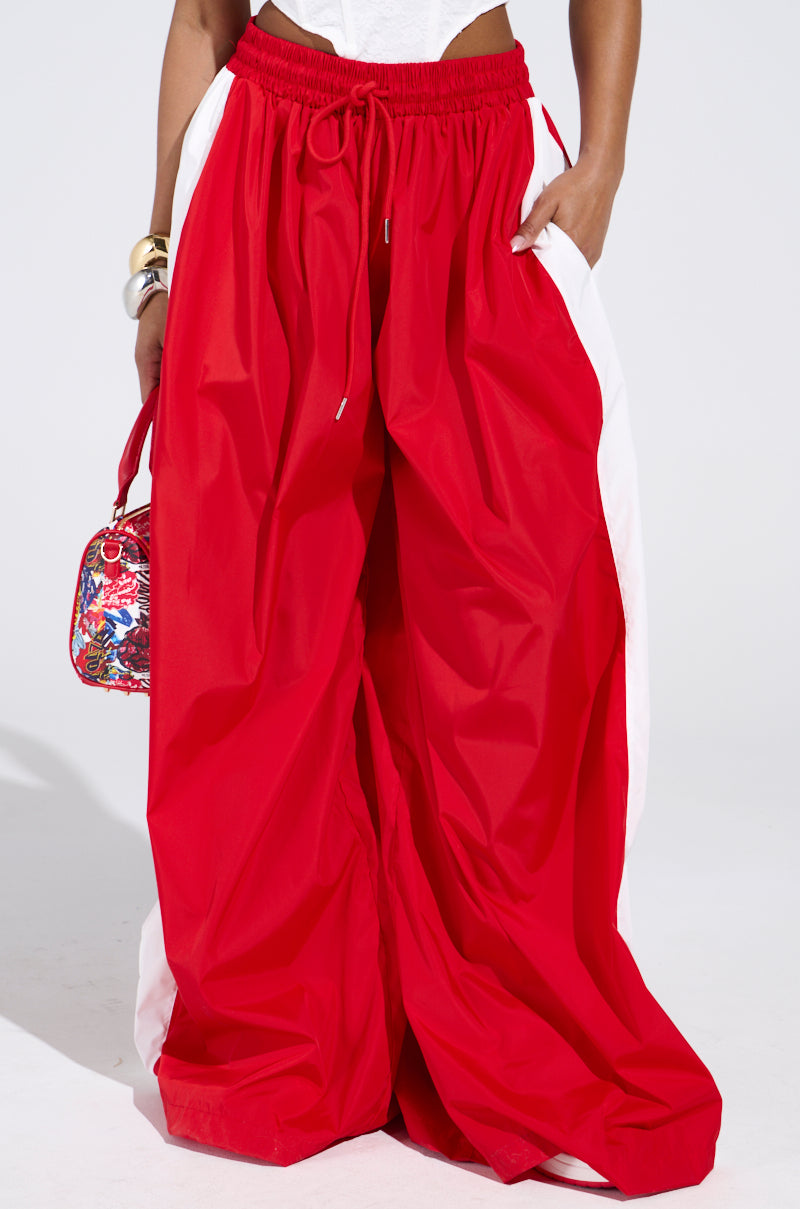 A person wearing the BREAKING THE RULES TRACK PANT, which features a high-rise fit, bright red color, wide leg silhouette, and white side panels, accessorized with a colorful handbag and gold-and-white bracelets.