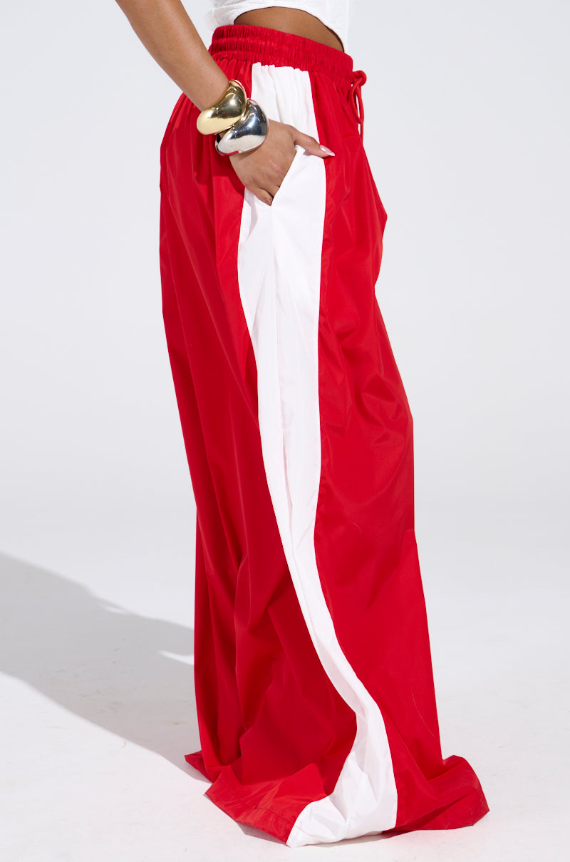 Someone wears the BREAKING THE RULES TRACK PANT—loose red with a wide white side stripe and wide leg silhouette—paired with a sleeveless top and chunky metallic bangles, standing against a plain white background.