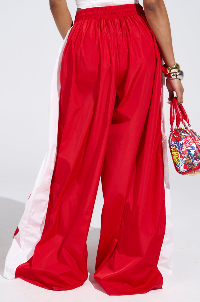 A person wears BREAKING THE RULES TRACK PANT—high-rise, wide-leg red track pants with white side stripes—styled with a colorful mini handbag and chunky gold bracelets against a plain, light background.