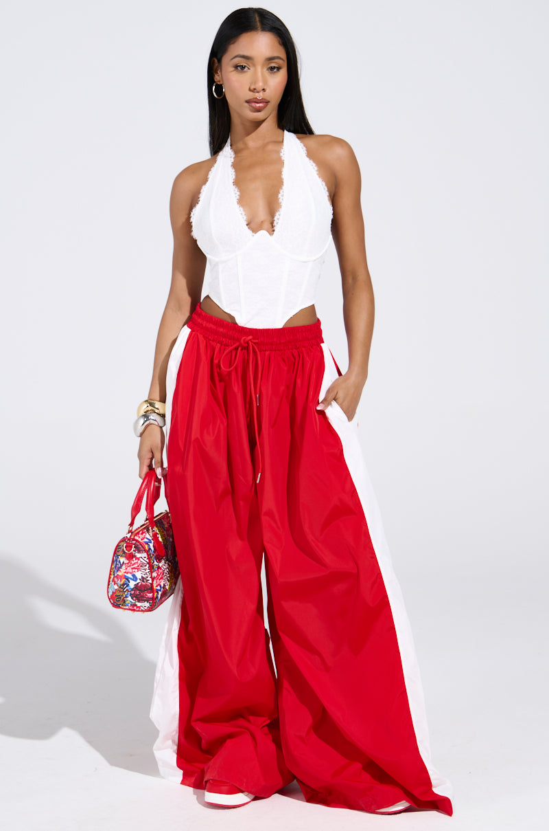 A woman poses in a white lace halter top and BREAKING THE RULES TRACK PANT—red, high-rise, wide-leg with white side panels—paired with a colorful handbag, bracelets, and matching white and red sneakers.