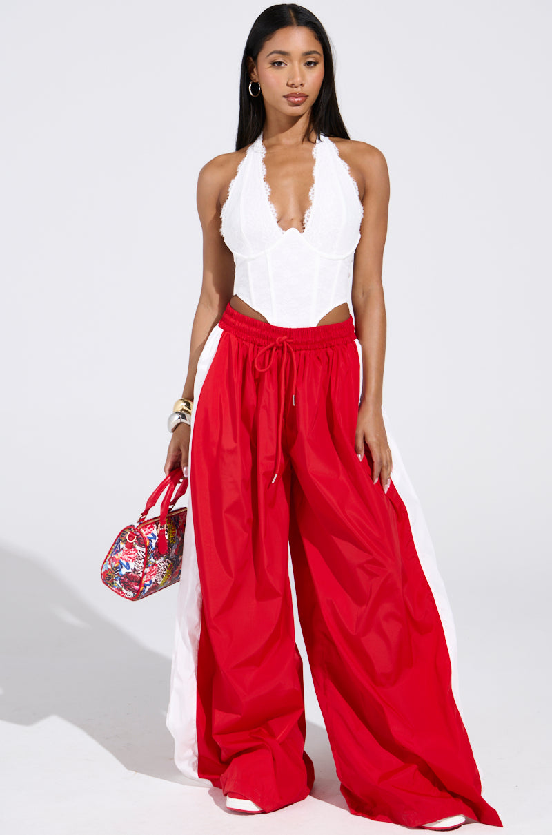 A woman stands against a plain white background wearing the BREAKING THE RULES TRACK PANT in red with white side panels, paired with a white halter lace top and holding a colorful patterned handbag.