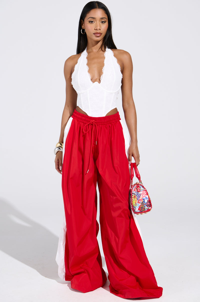 A woman stands confidently in the BREAKING THE RULES TRACK PANT with a wide leg silhouette, paired with a white lace halter top. She holds a colorful small handbag and accessorizes with hoop earrings and bangles.