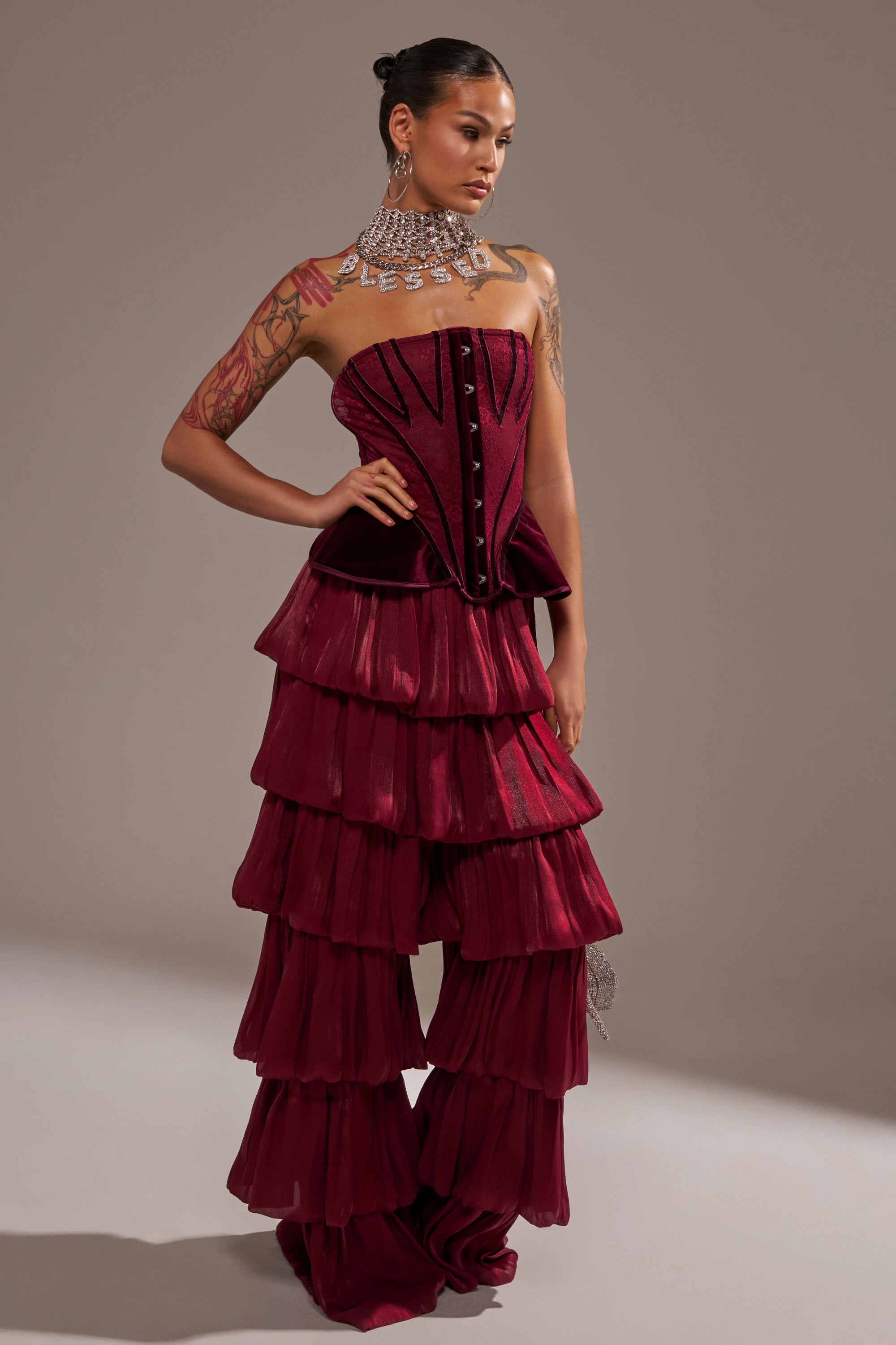 A person models the STAR OF THE NIGHT FLOUNCY PANT IN BURGUNDY, featuring high-rise, tiered ruffle wide legs. They pair it with a strapless burgundy corset, bold statement necklace, and show off arm tattoos against a plain background.