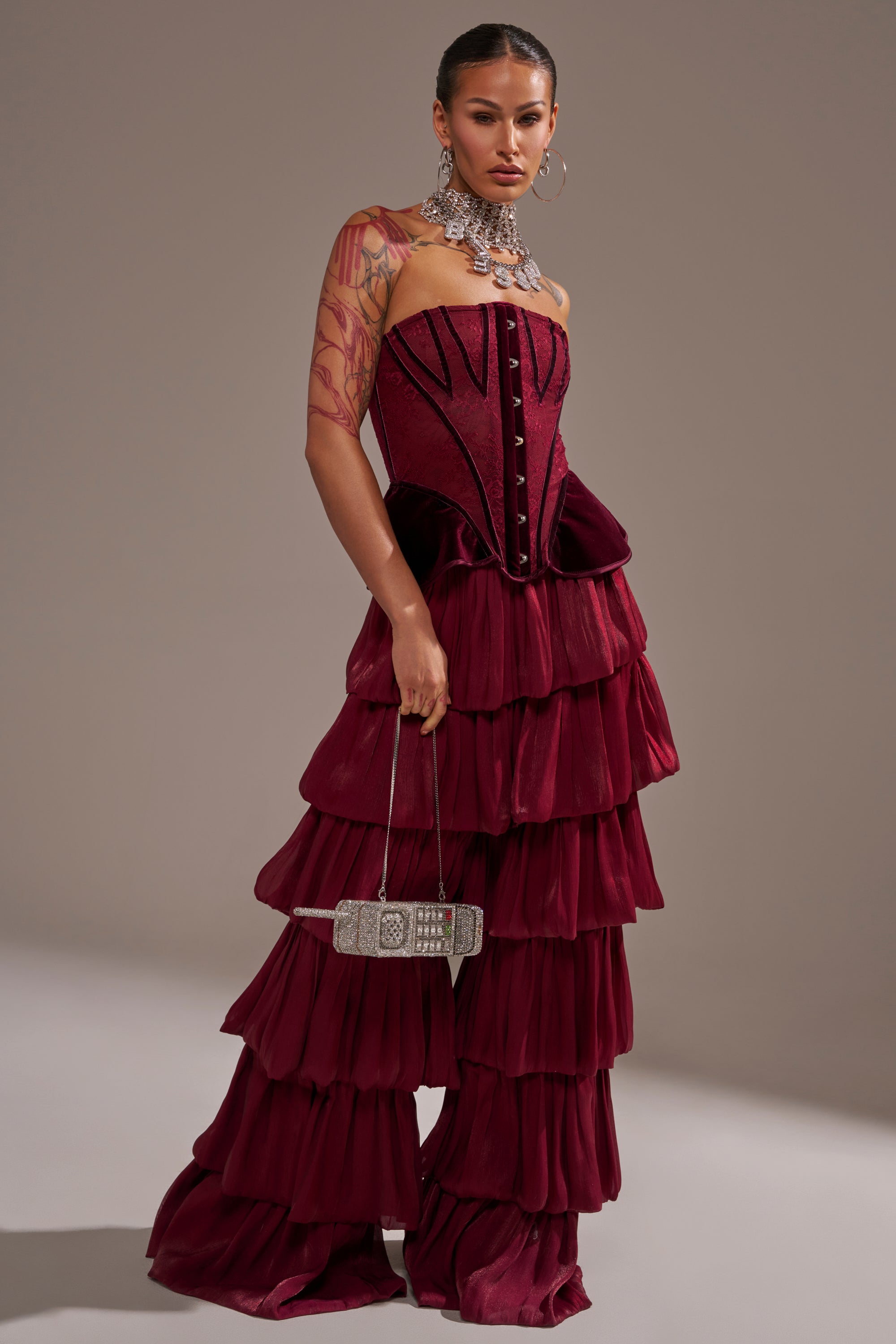 A woman poses in the STAR OF THE NIGHT FLOUNCY PANT IN BURGUNDY, paired with a chunky jeweled necklace. She shows tattoos on her arm and holds a rectangular silver clutch. 1