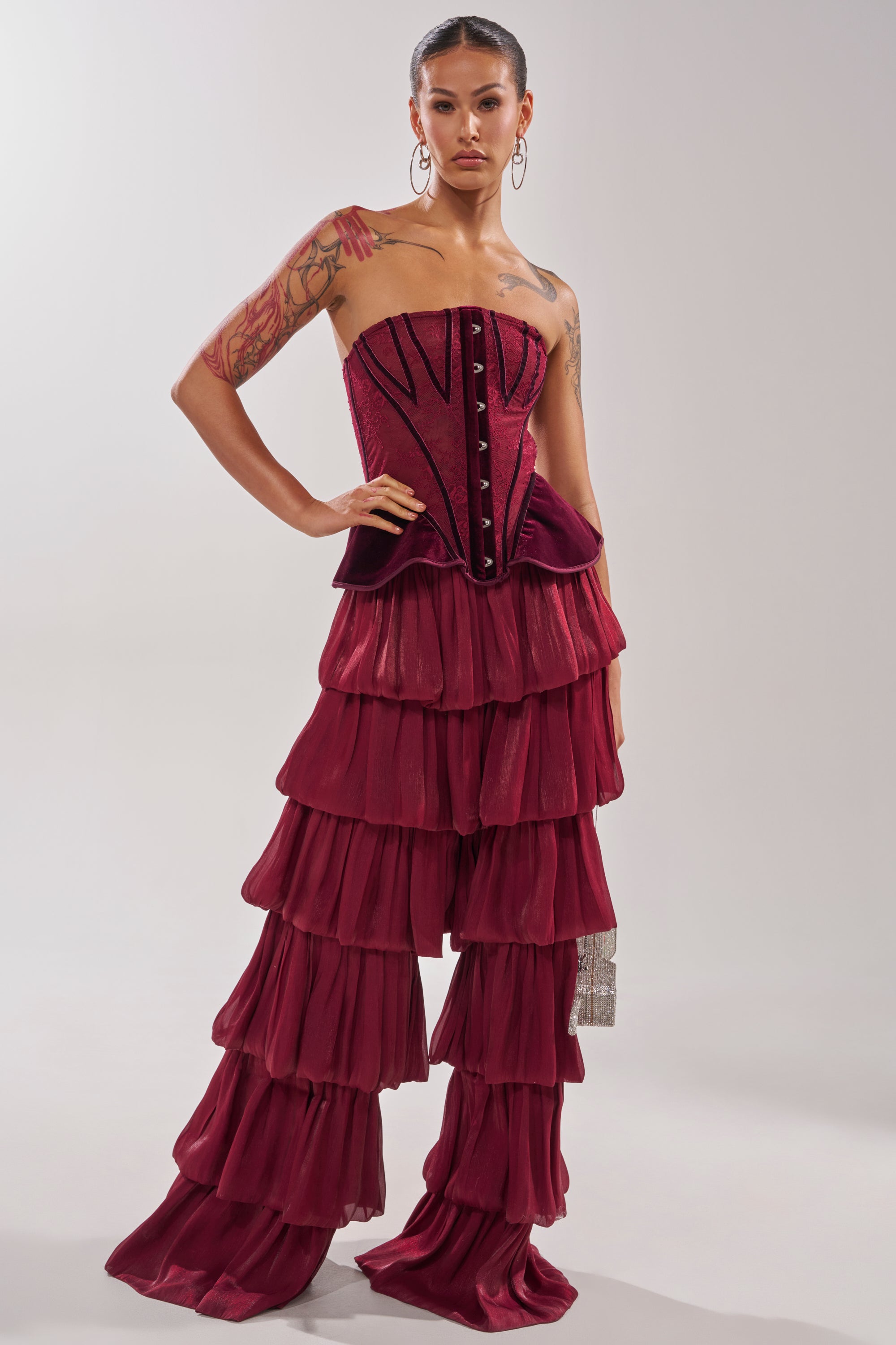 A woman poses confidently in the STAR OF THE NIGHT FLOUNCY PANT IN BURGUNDY, high-rise ruffle wide-leg pants with a chiffon-like fabric. She accessorizes with hoop earrings, shows arm tattoos, and stands against a plain light background. 2