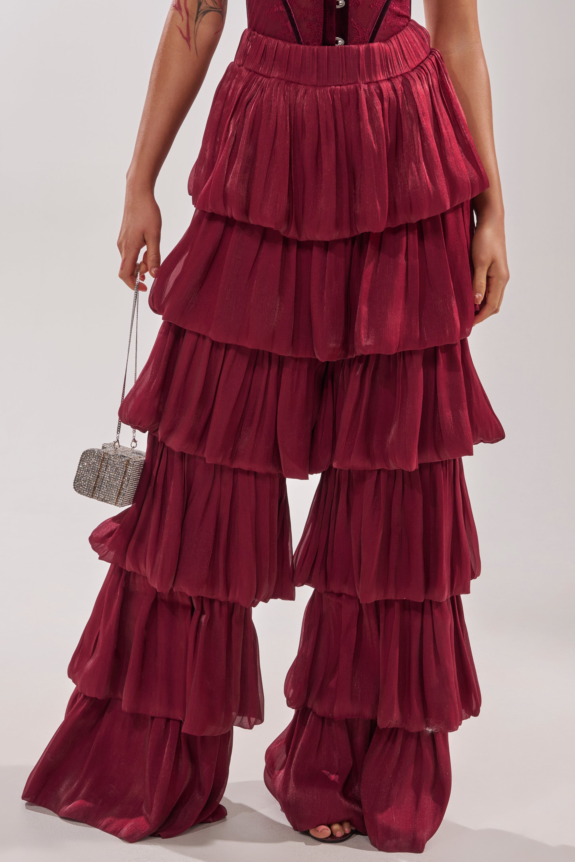 A person wearing the STAR OF THE NIGHT FLOUNCY PANT IN BURGUNDY holds a small, sparkly handbag. Only their lower body is shown against a plain background. 3