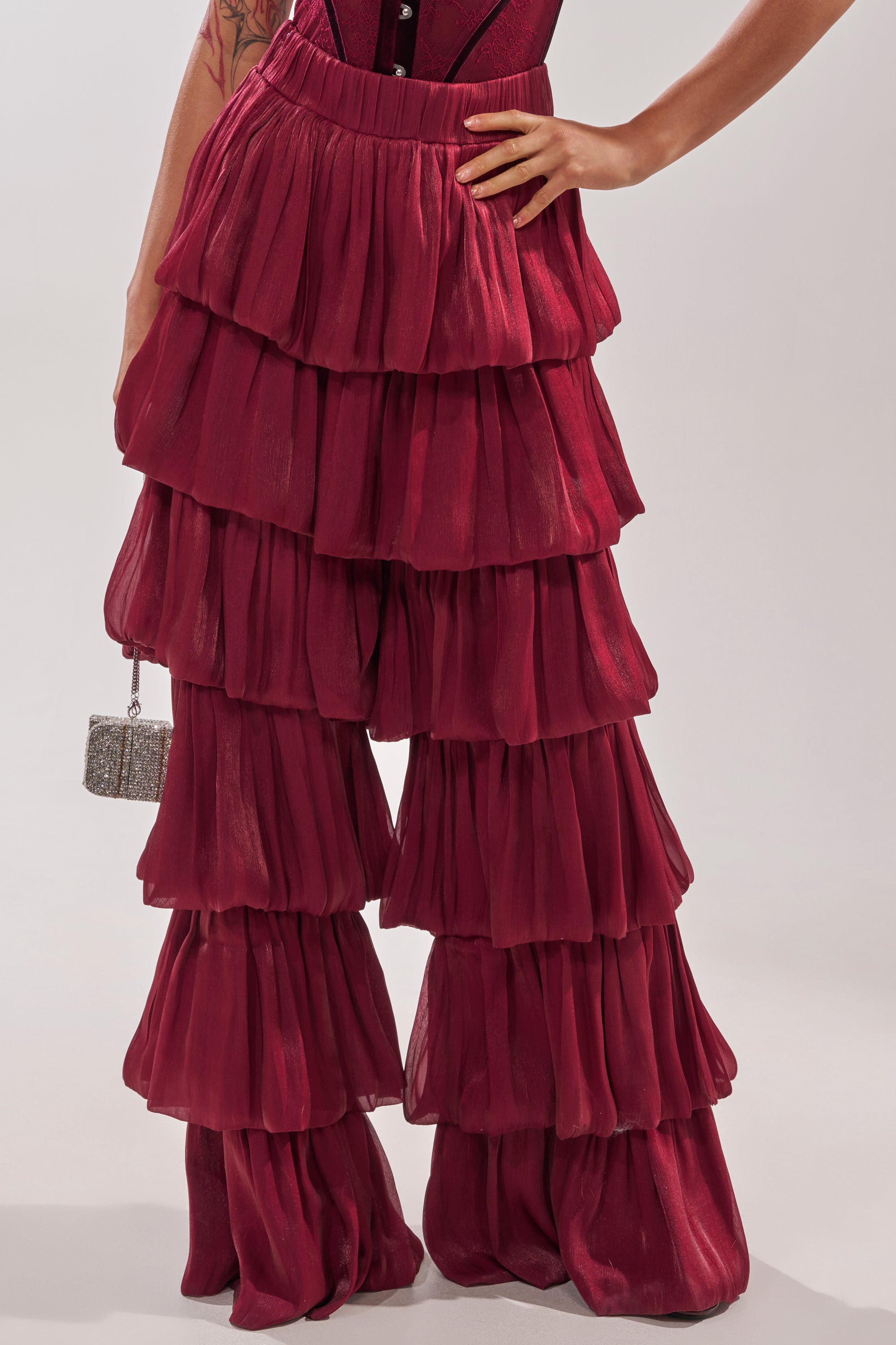 A person wears the STAR OF THE NIGHT FLOUNCY PANT IN BURGUNDY, featuring a high-rise fit and tiered ruffles in sheer fabric, holding a small silver box-shaped handbag. The focus is on the wide-leg pants and their hand. 4