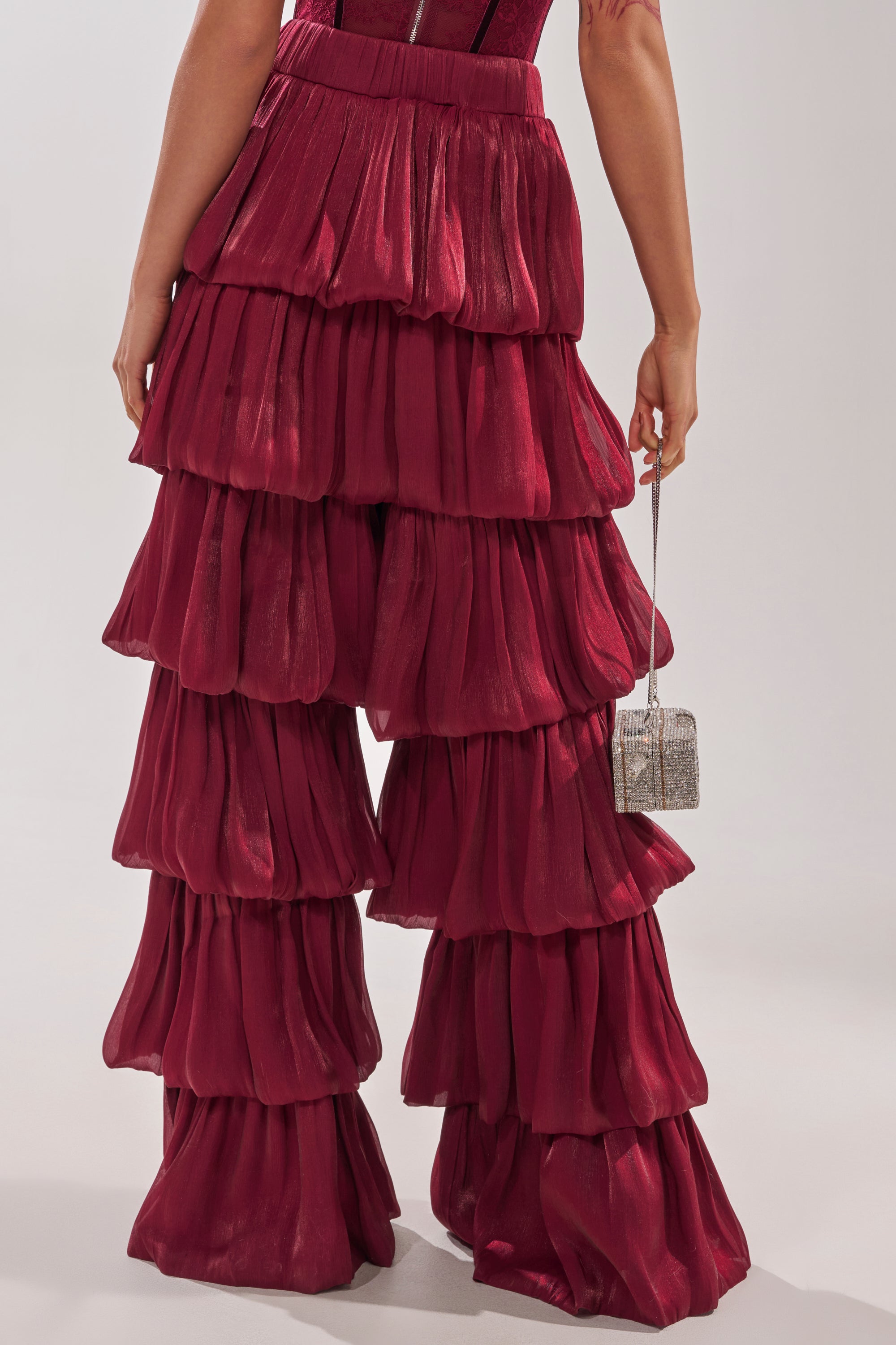 A person wears the STAR OF THE NIGHT FLOUNCY PANT IN BURGUNDY—deep red, wide-leg and shiny. They hold a small silver box purse, and the bold high-rise pants stand out against a plain light gray background. 6