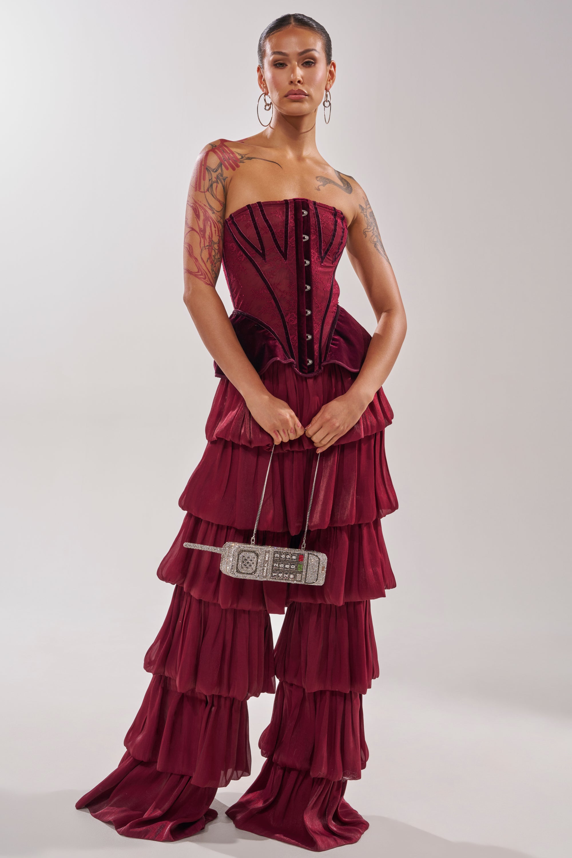 A person stands against a plain background wearing the STAR OF THE NIGHT FLOUNCY PANT IN BURGUNDY, featuring ruffled wide-leg chiffon-like pants, hoop earrings, visible tattoos, and holding a small retro cellphone-shaped purse. 7