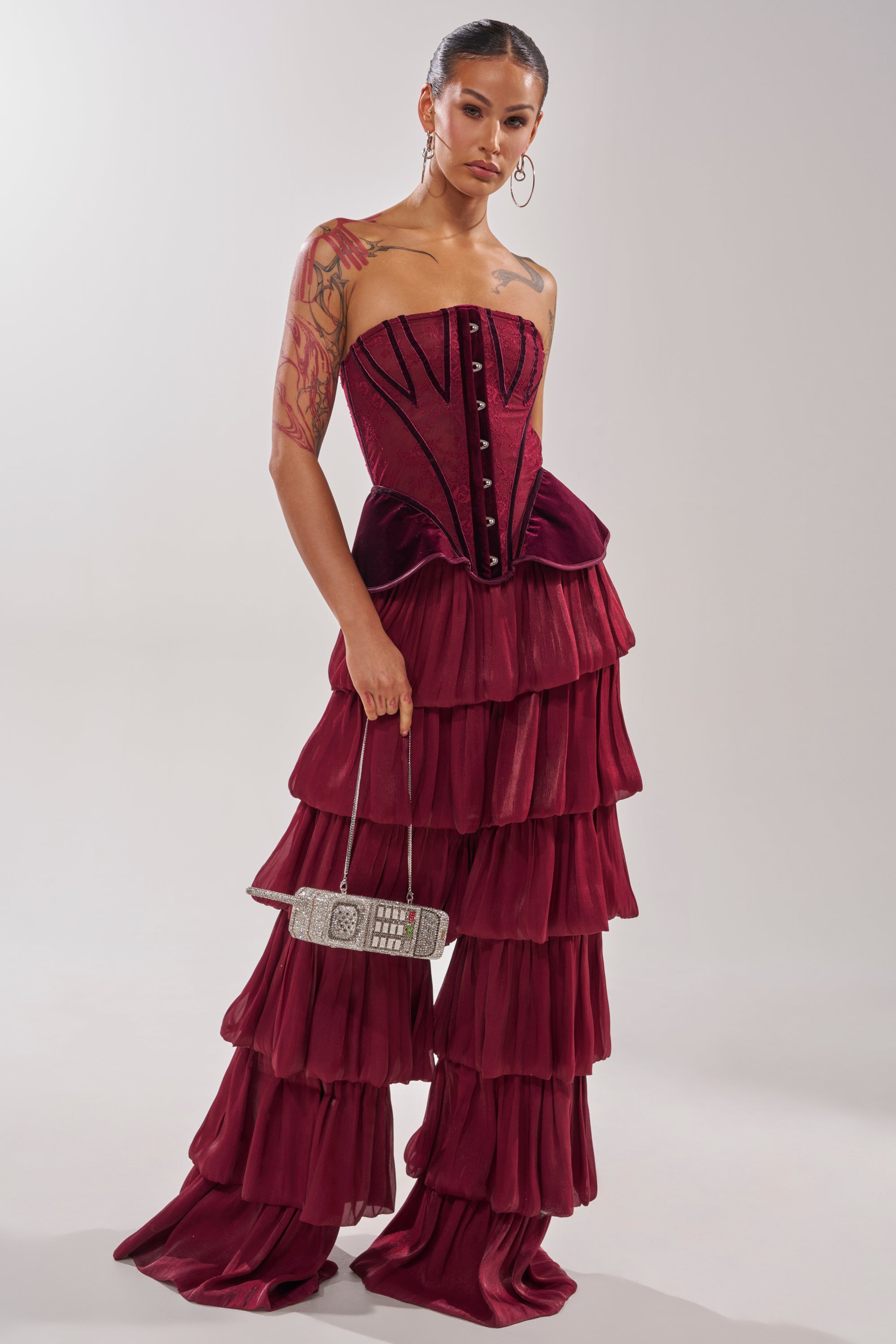 A woman in STAR OF THE NIGHT FLOUNCY PANT IN BURGUNDY and a strapless corset top holds a small decorative handbag, with slicked-back hair, large hoop earrings, and an arm tattoo. 8